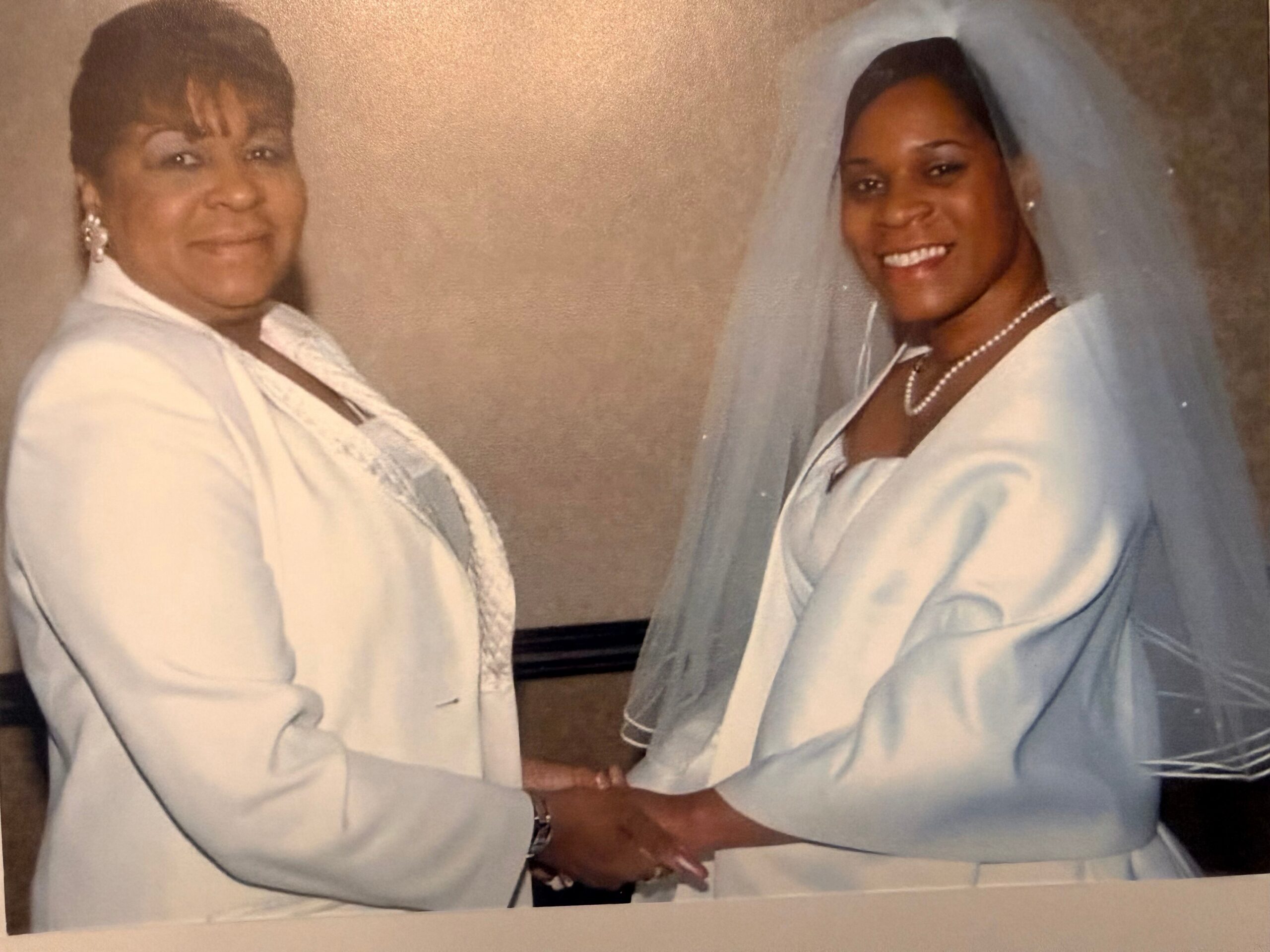 Bride in a white gown and veil greets a woman in a white outfit; they clasp hands and smile for a wedding photo.