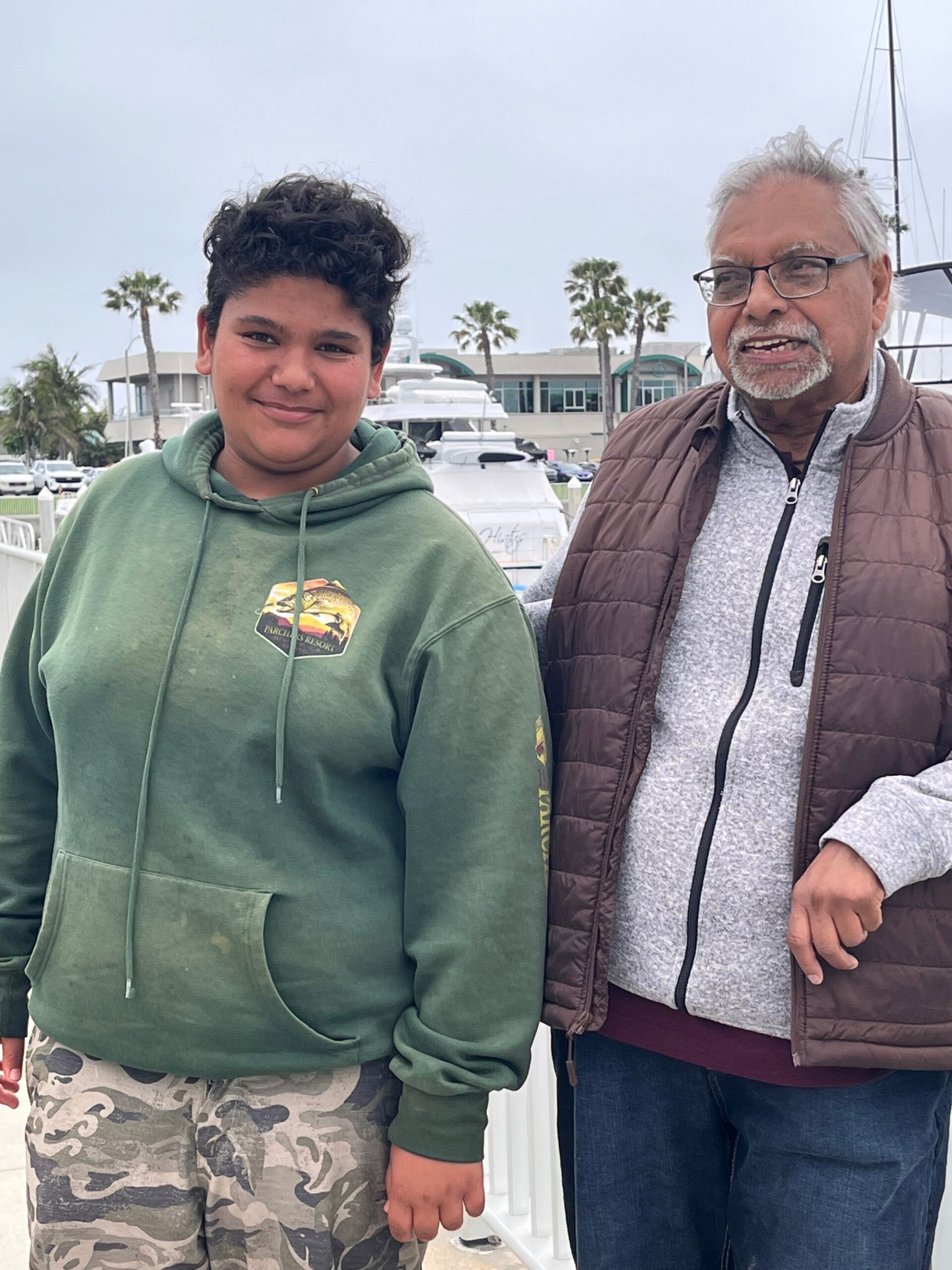 Two people posing together outdoors at a marina with palm trees and boats in the background: a young person in a green hoodie on the left and an older man with glasses in a brown vest on the right.