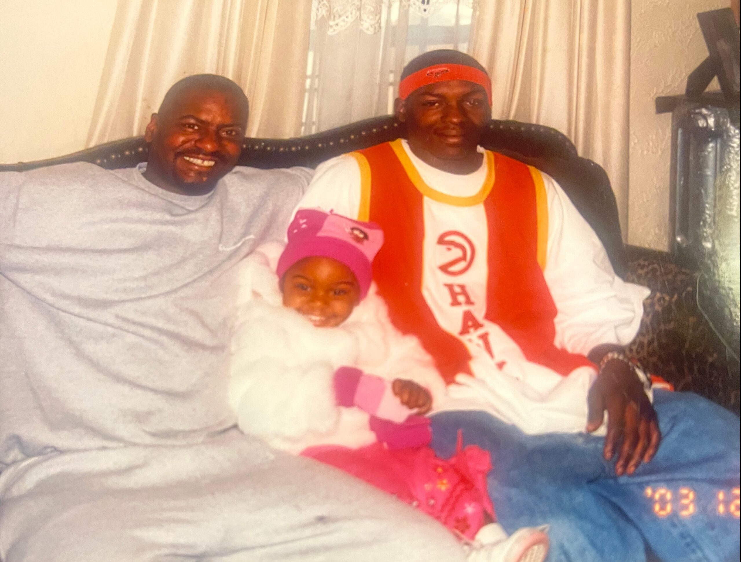 Three people pose on a sofa: a man in gray with a friendly smile, another man in a red-and-yellow jersey, and a child in a pink hat, all looking at the camera.