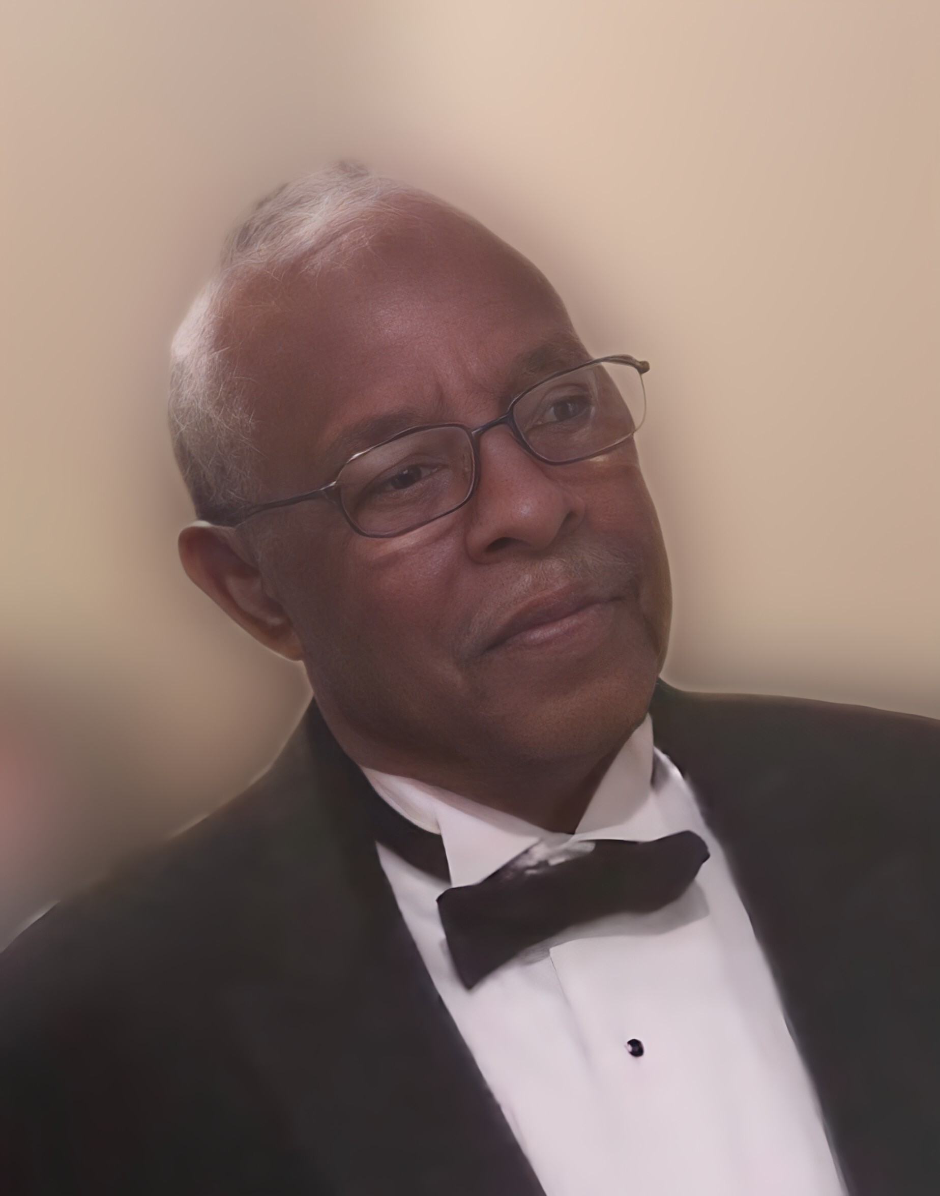 Portrait of a man in a black tuxedo and bow tie, looking to his right against a soft beige background, wearing glasses.