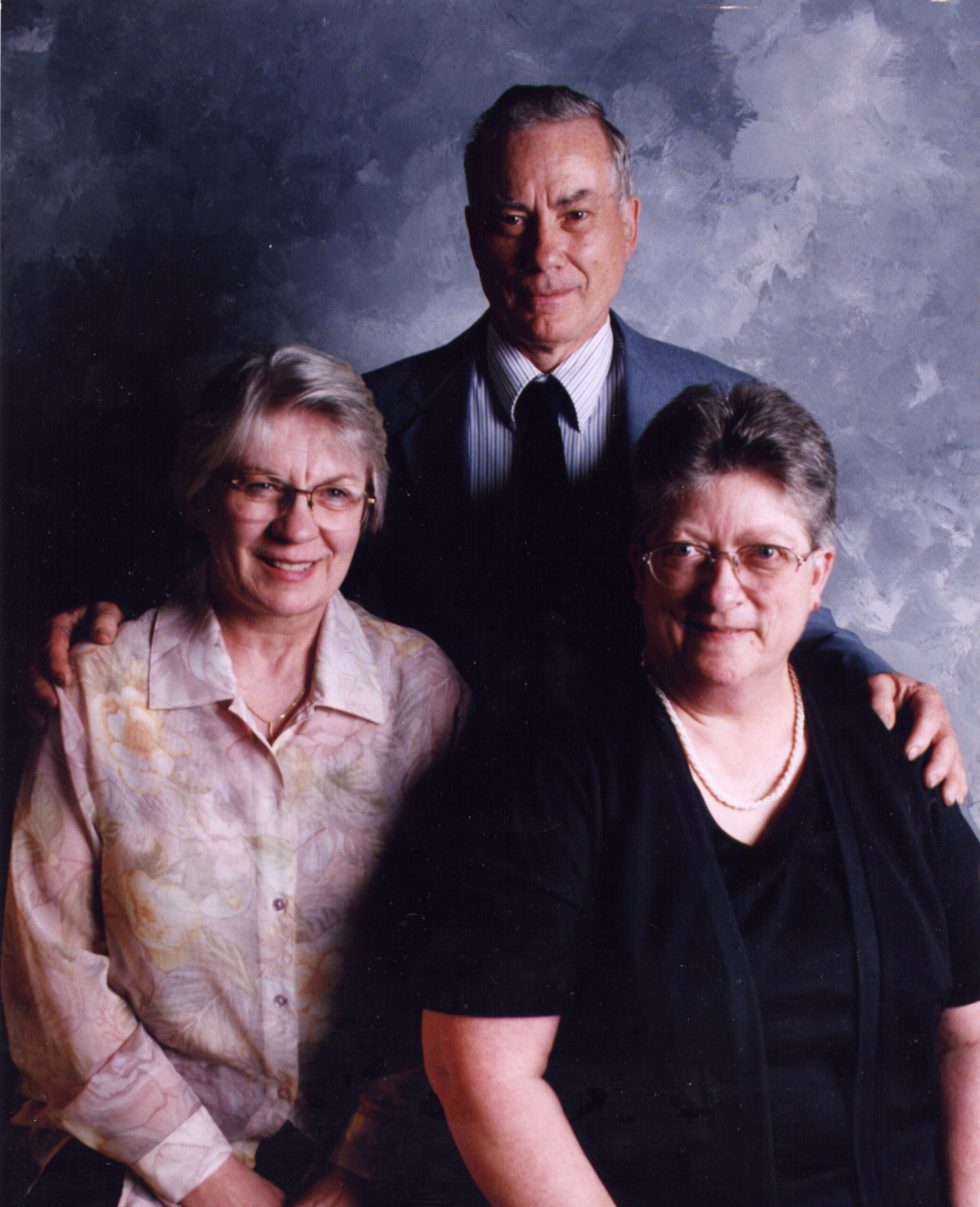 Three adults pose together for a family portrait: a man in a suit behind two women, their arms around each other, smiling at the camera.