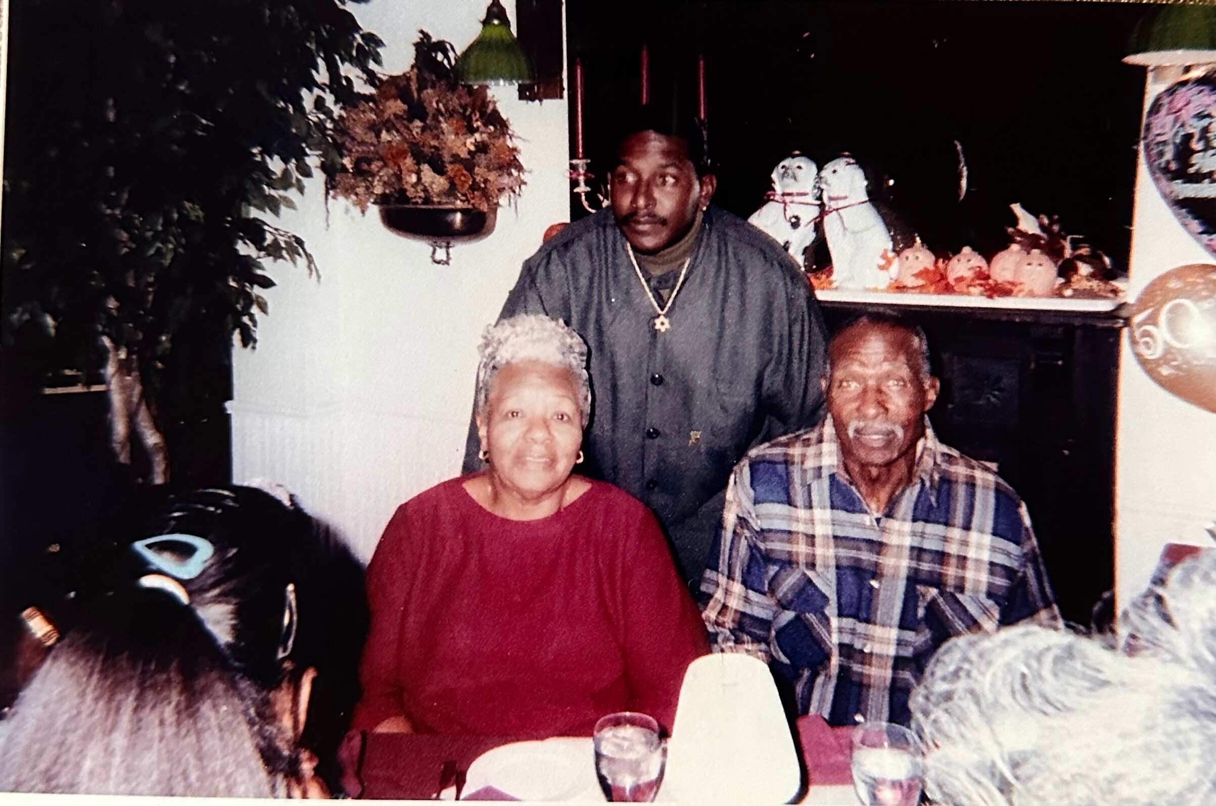 A smiling elderly woman in a red top and a man in a plaid shirt sit at a dining table, with a younger man standing behind them, in a warmly dressed family gathering with autumn decor in the background.