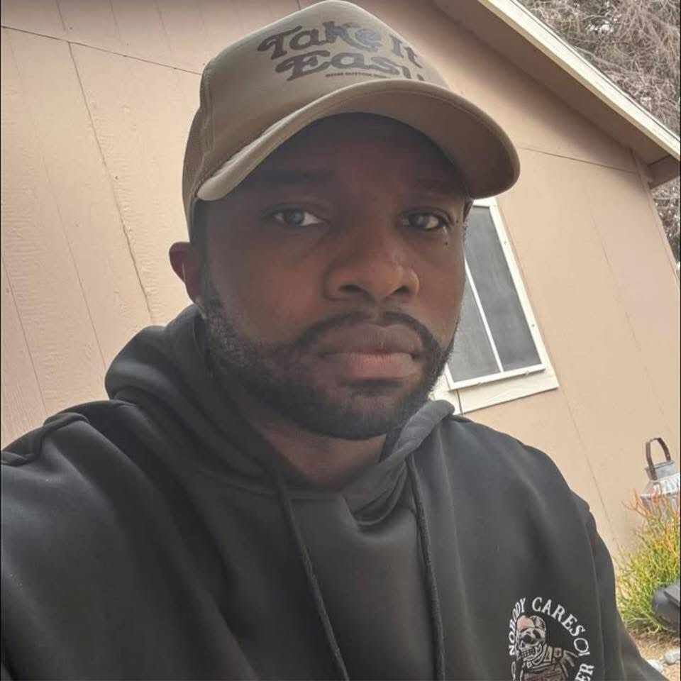 Close-up of a man wearing a beige baseball cap and black hoodie, standing outside a tan house with a window behind him.