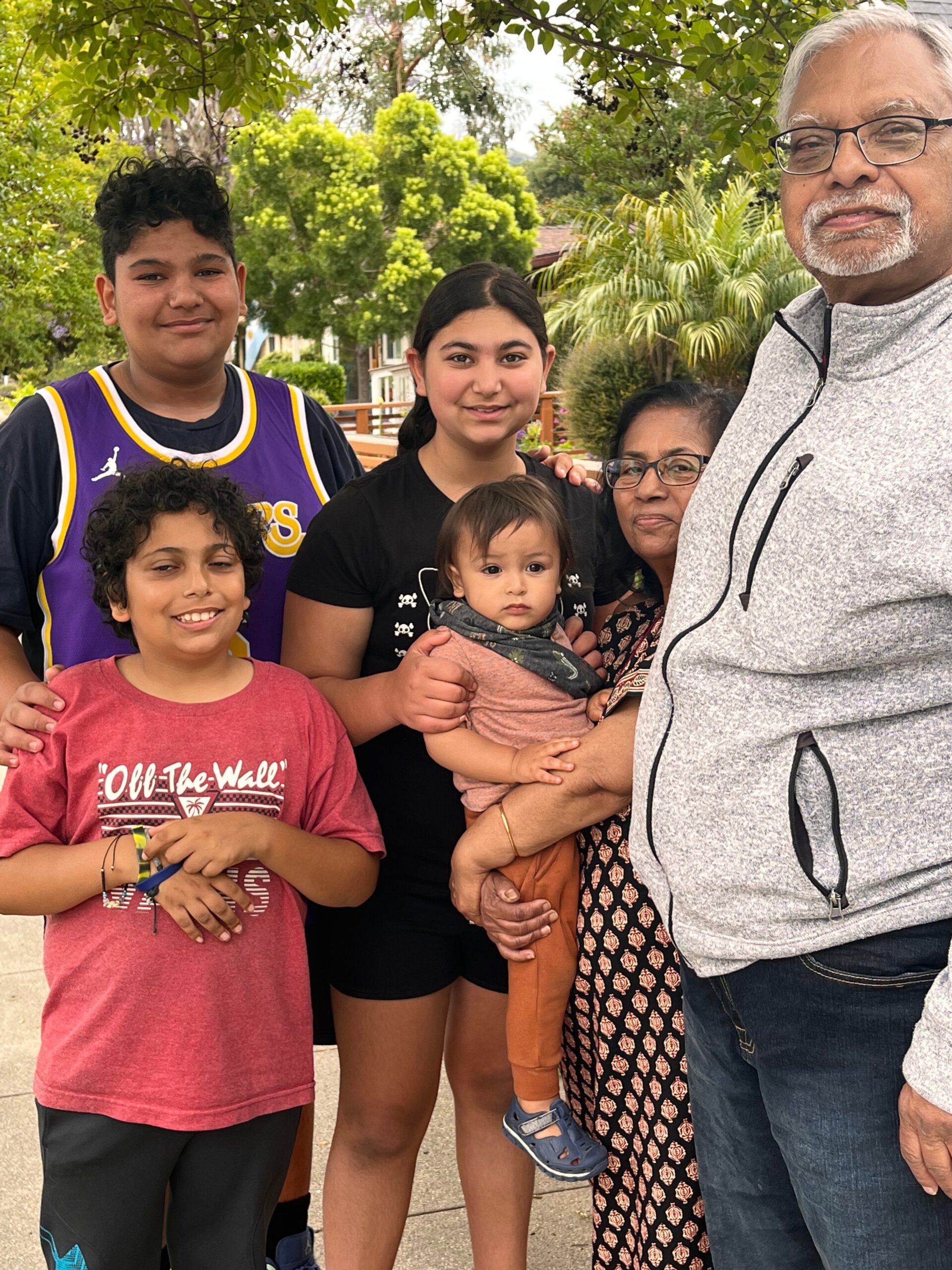 Multigenerational family posing outdoors among trees and shrubs, smiling at the camera.