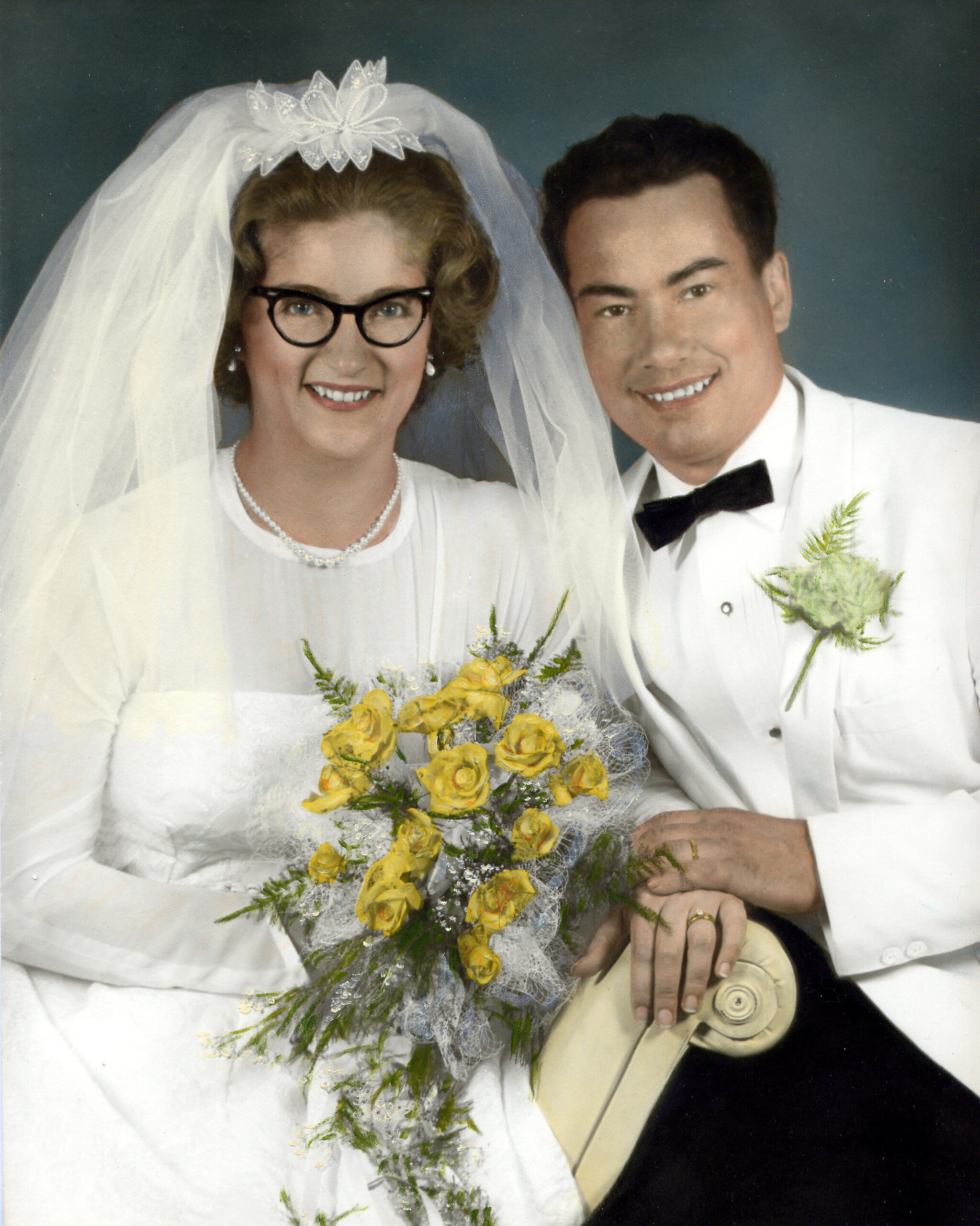 Bride and groom pose in a wedding portrait; she wears a veil, glasses, and a pearl necklace, holding a yellow rose bouquet beside him in a white suit.