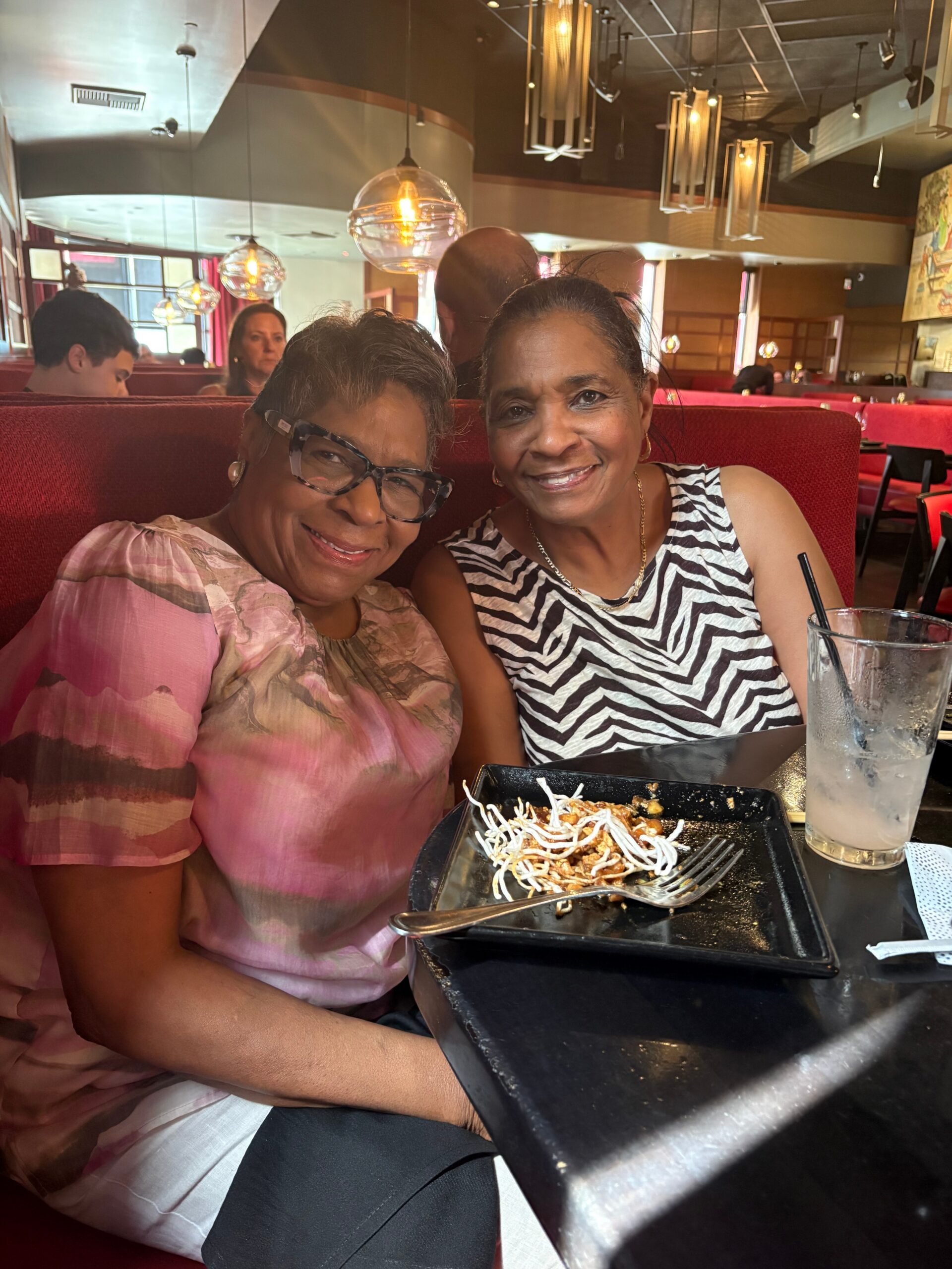 Two women sit close together at a red booth in a restaurant, smiling at the camera with a plate of noodles in front of them and a tall glass with a straw on the table.