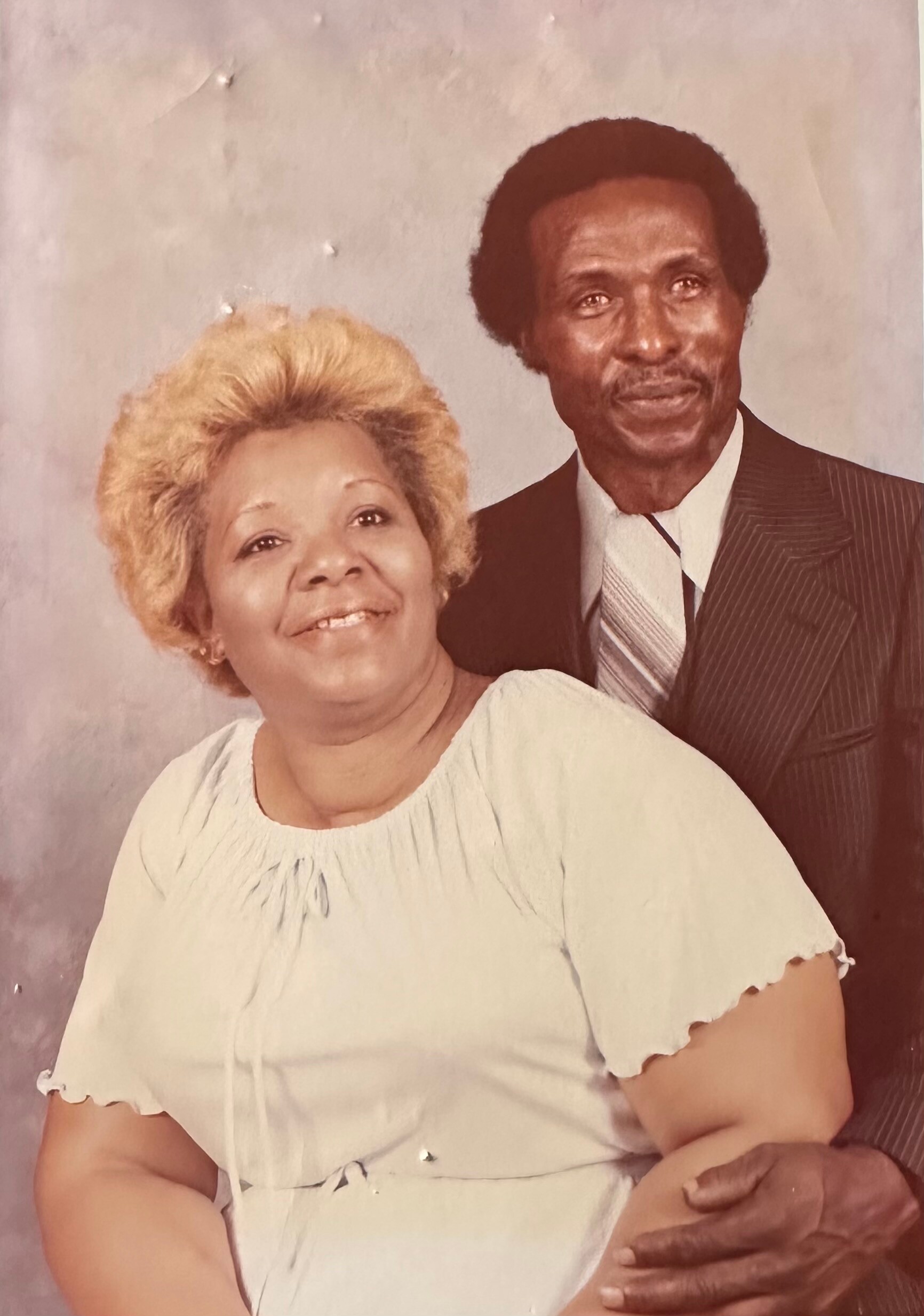 Smiling elderly woman in a light blouse beside an older man in a suit posing for a studio portrait with a gentle embrace.
