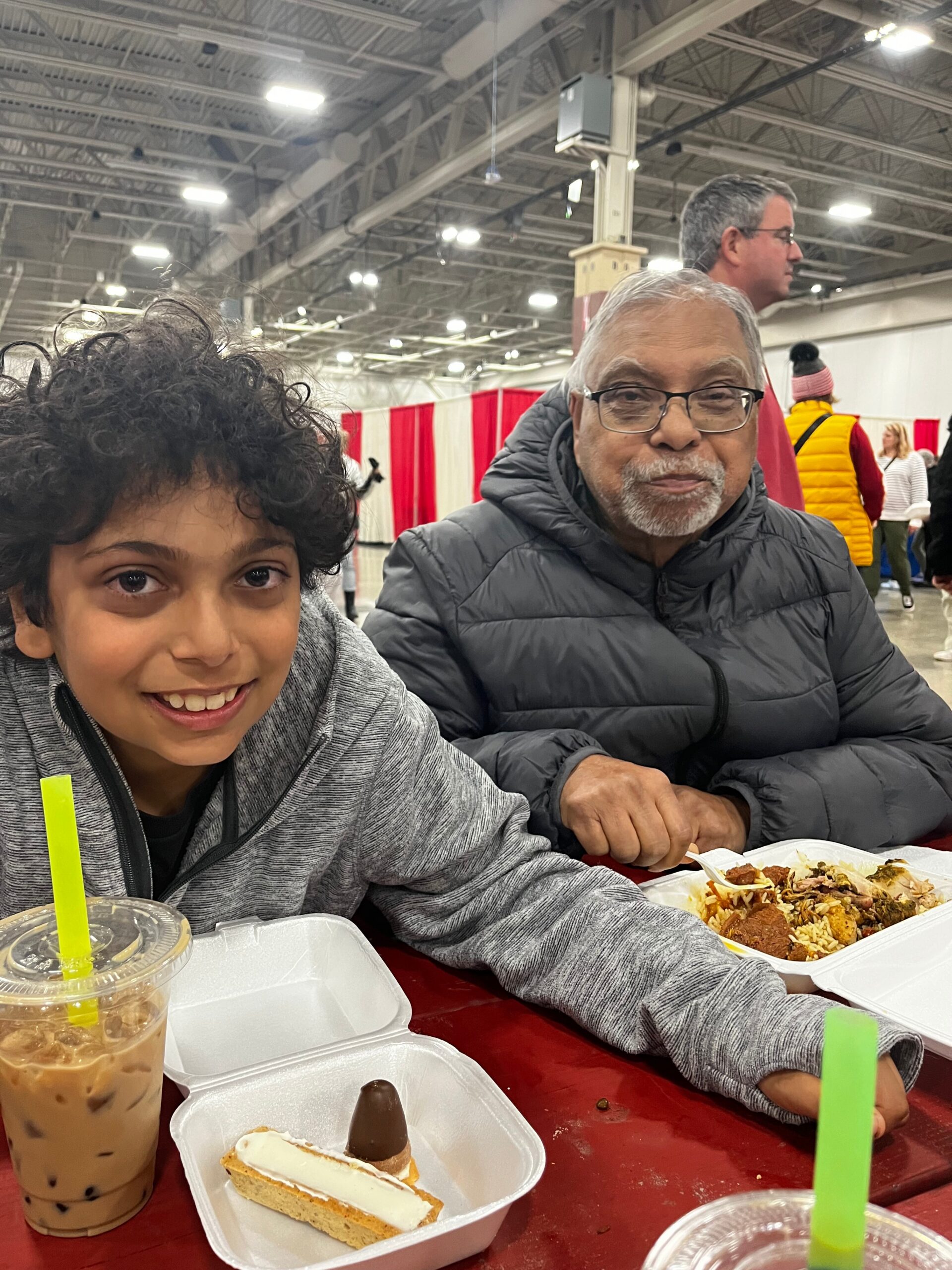 Young boy and an older man with glasses sit at a red table in a large indoor event hall, enjoying dessert and drinks together.