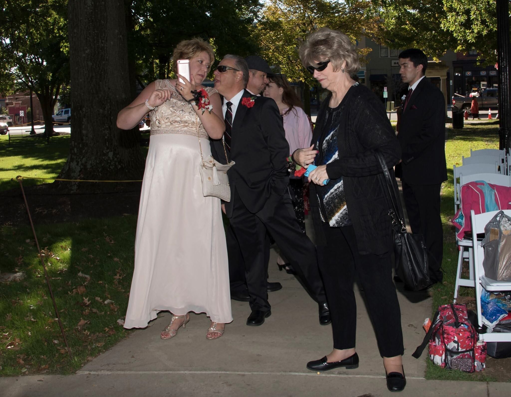 Group of formally dressed people outdoors, a woman in a light dress takes a selfie with her phone while others chat nearby.