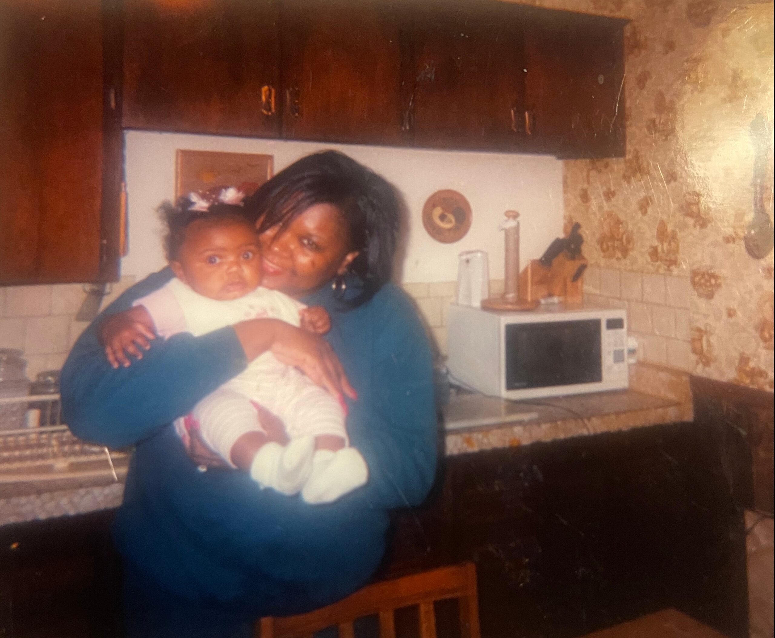 Mother holding a baby in a kitchen, both looking at the camera, with wooden cabinets and a microwave in the background.