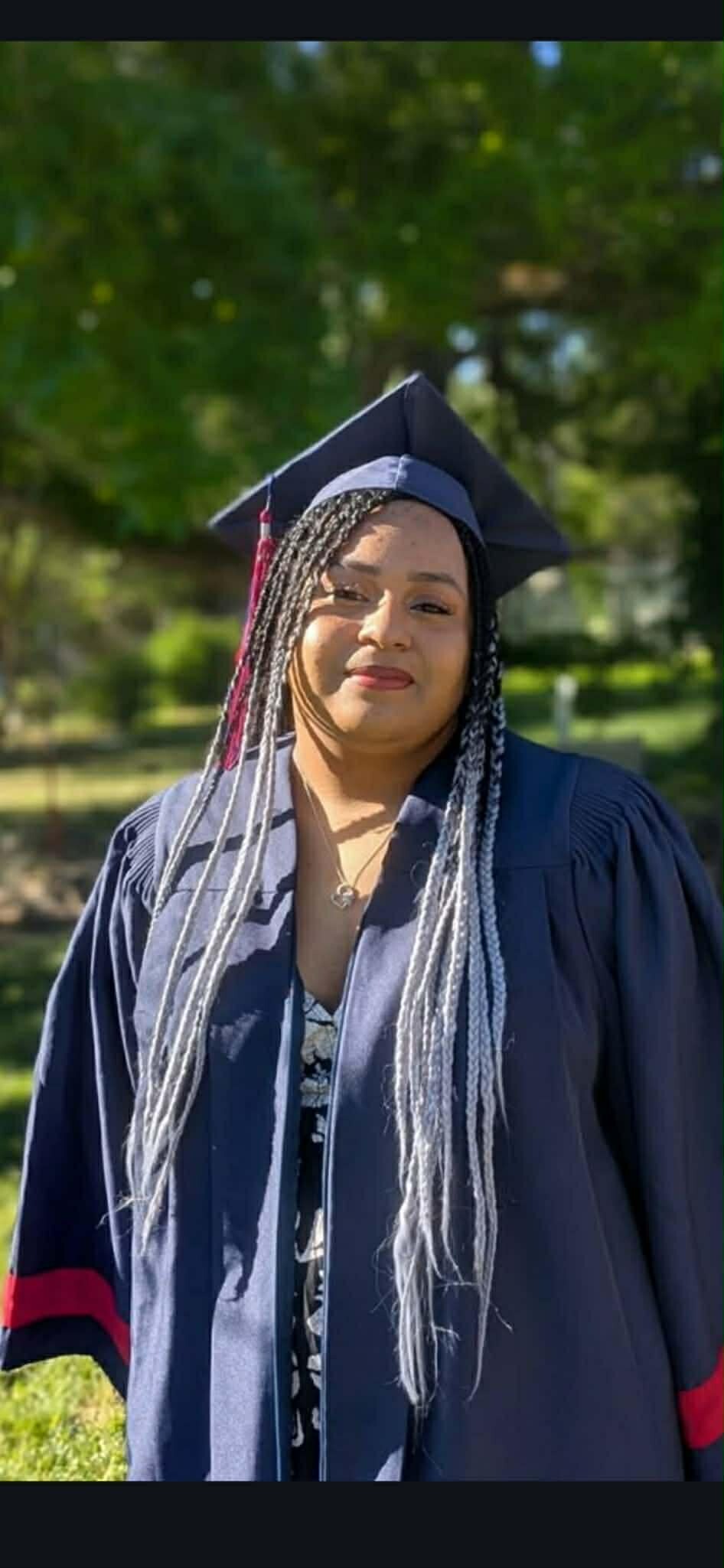 Graduate in a navy cap and gown with a red tassel, smiling outdoors in a park.