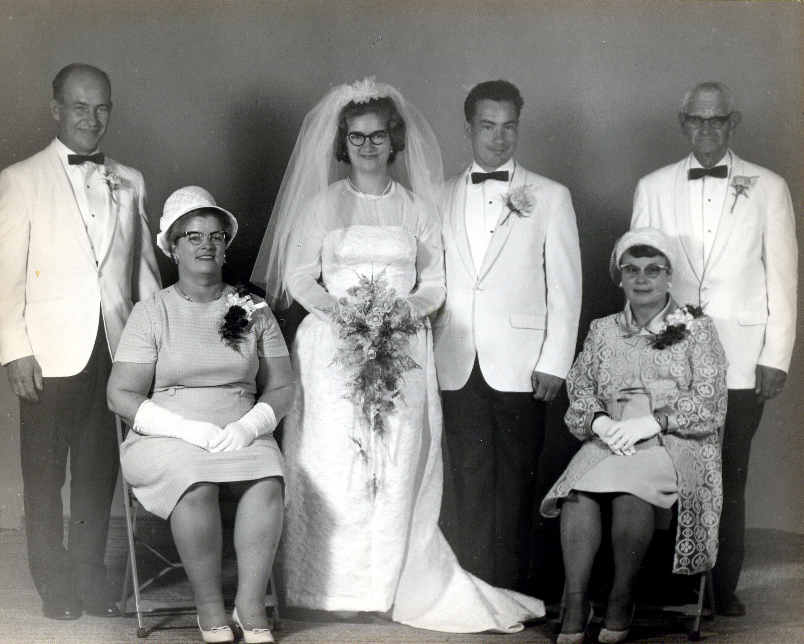 Black-and-white wedding portrait: bride in a white gown and veil center, flanked by groom in a white tuxedo and two older men in white jackets; two women seated at sides wearing hats and dresses.