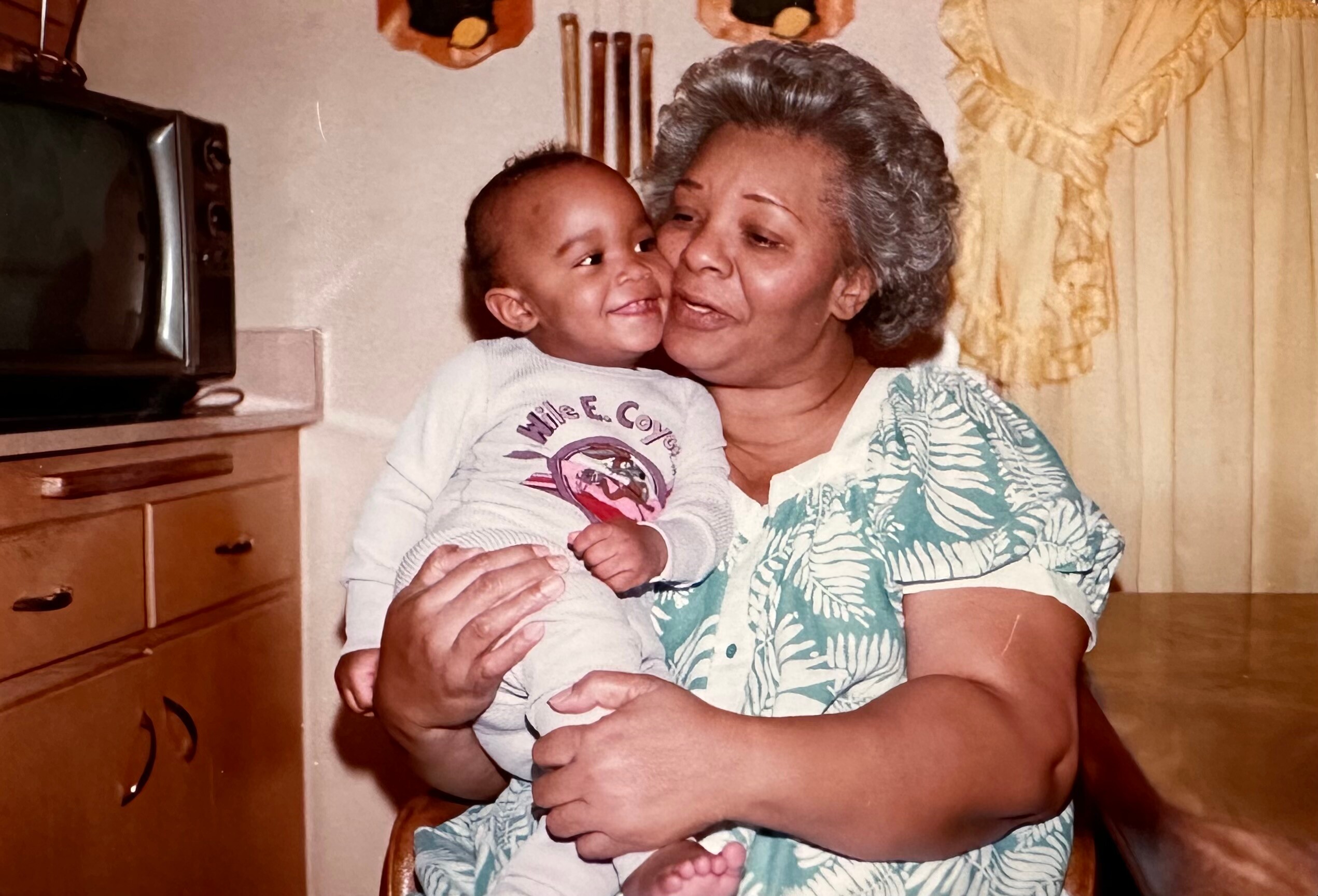 Grandmother cradling a smiling baby in a kitchen, both looking at each other warmly
