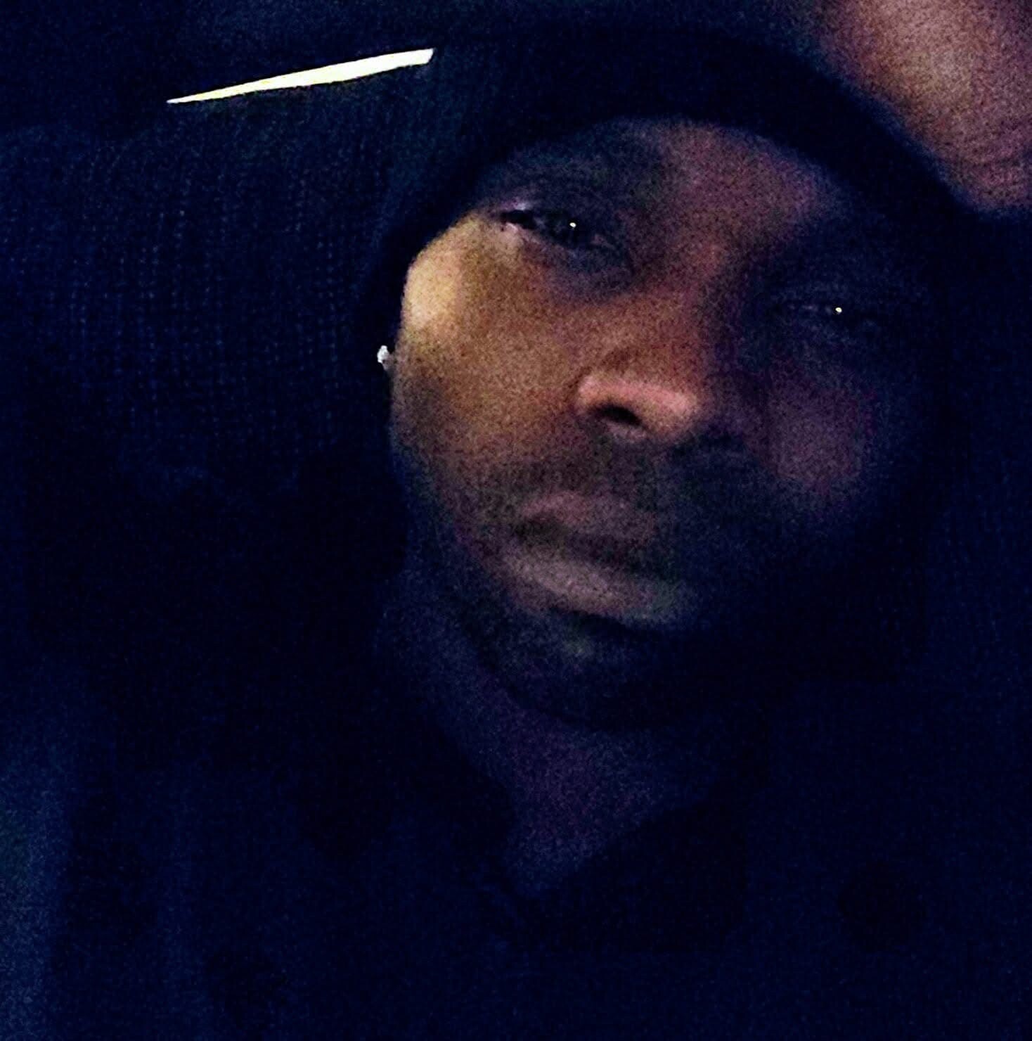 Close-up of a person wearing a dark hood and beanie in low light; only eyes and mouth are slightly visible.