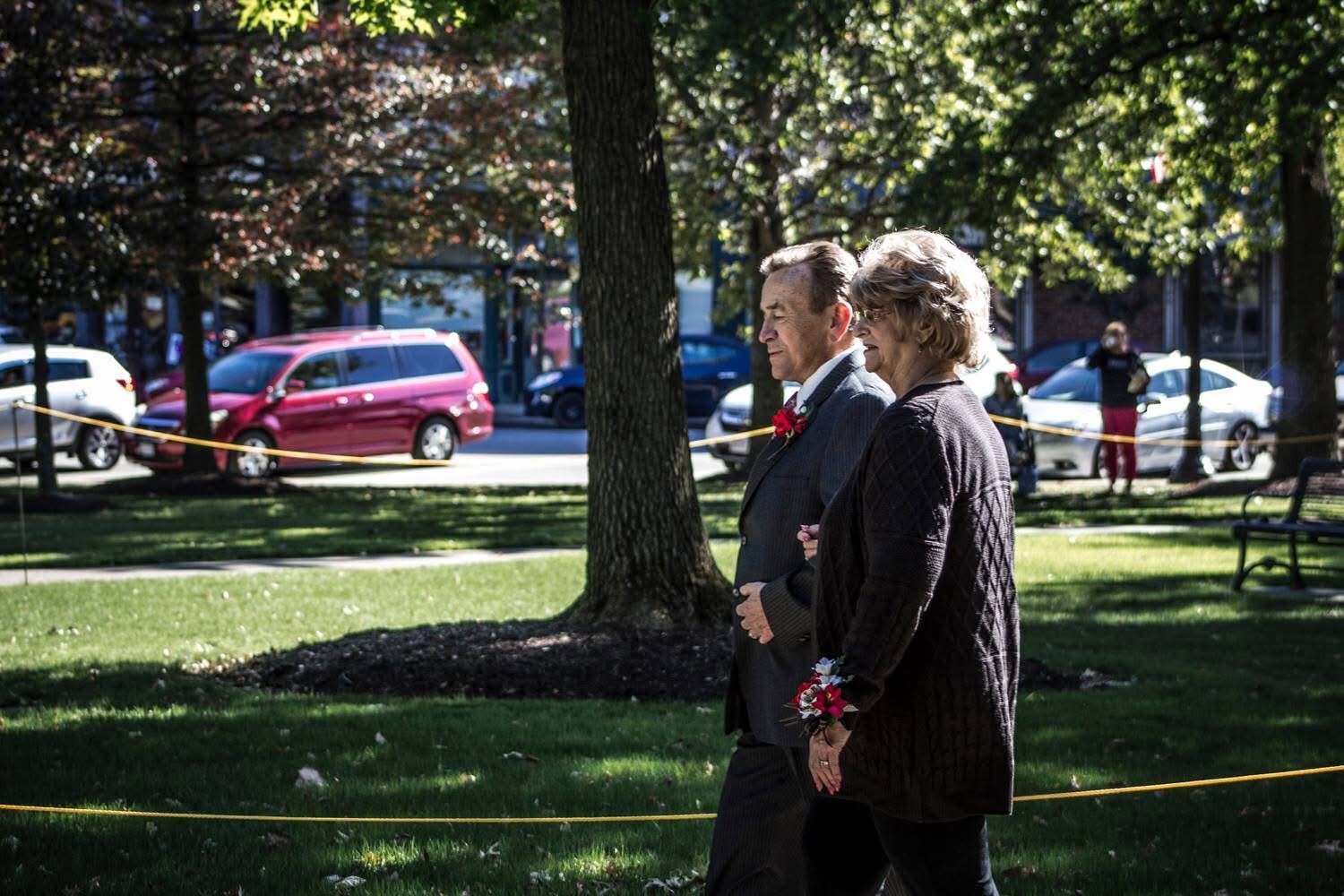 An elderly couple walks arm in arm through a sunlit park, wearing formal attire and red corsages, with cars and yellow tape in the background.