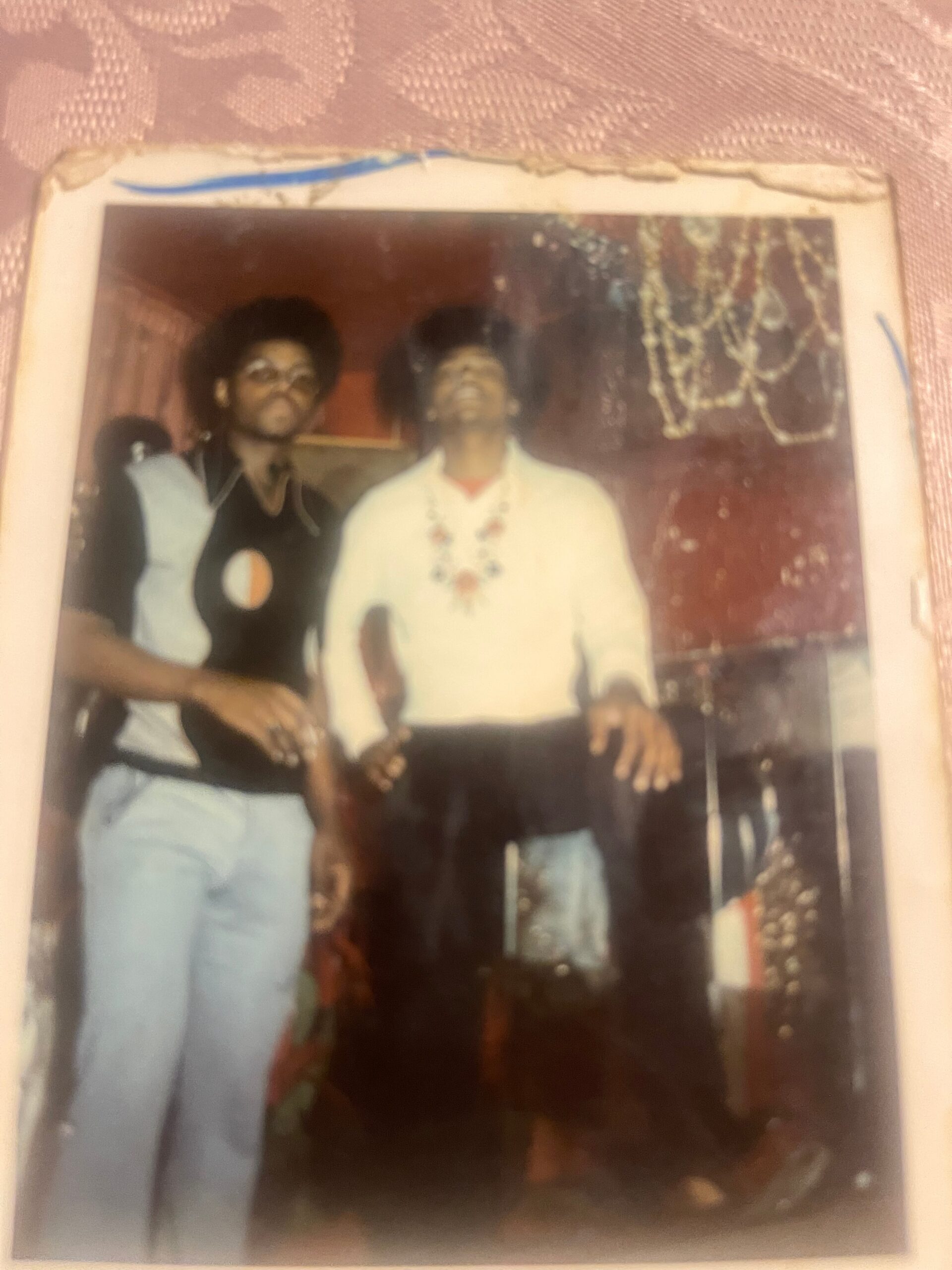 Three men with big afros posing together in a casual indoors photo, one in a white shirt with a bead necklace.