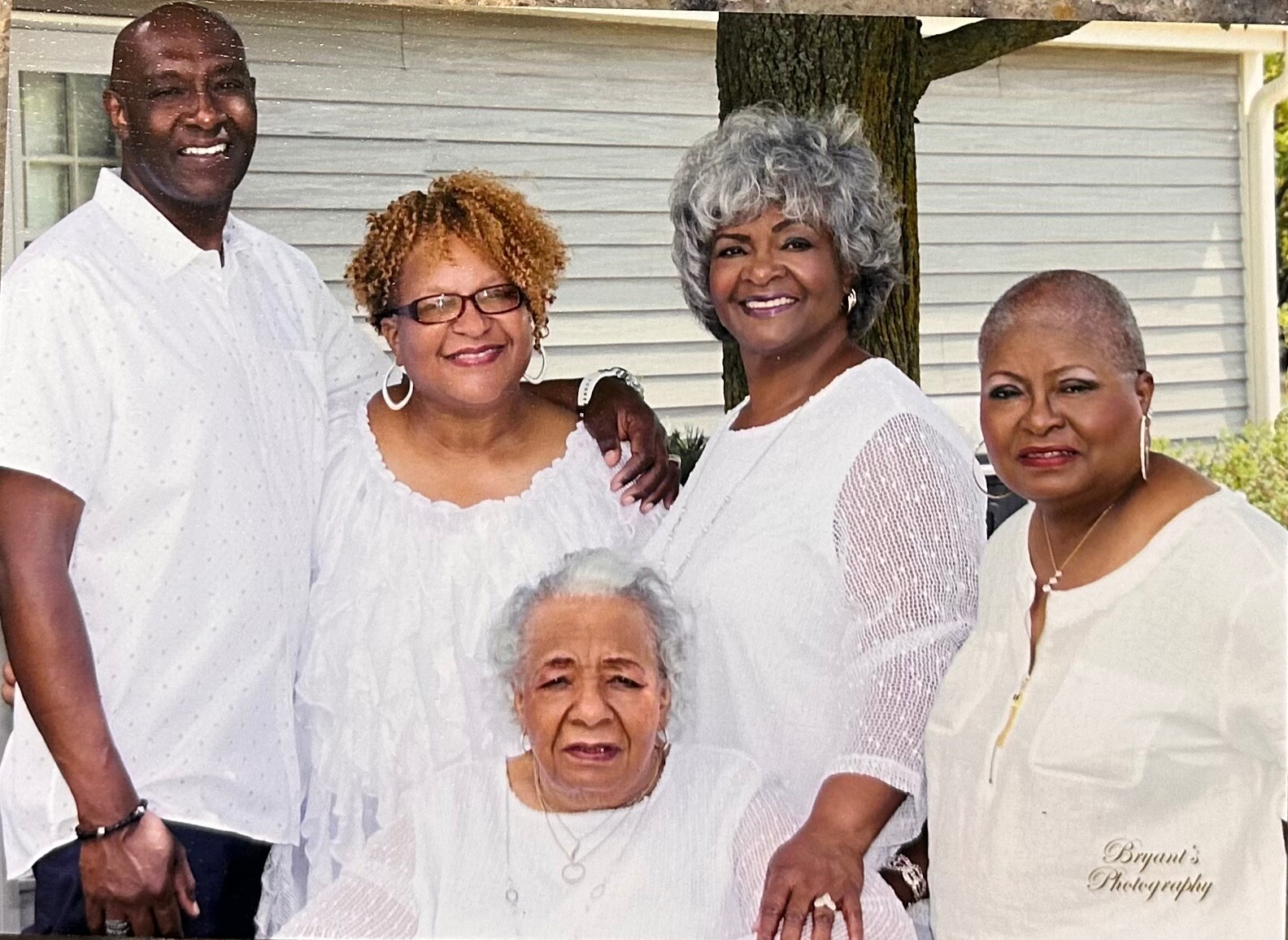 Group of five adults posing outdoors in white outfits, smiling for a family photo under a tree by a house.