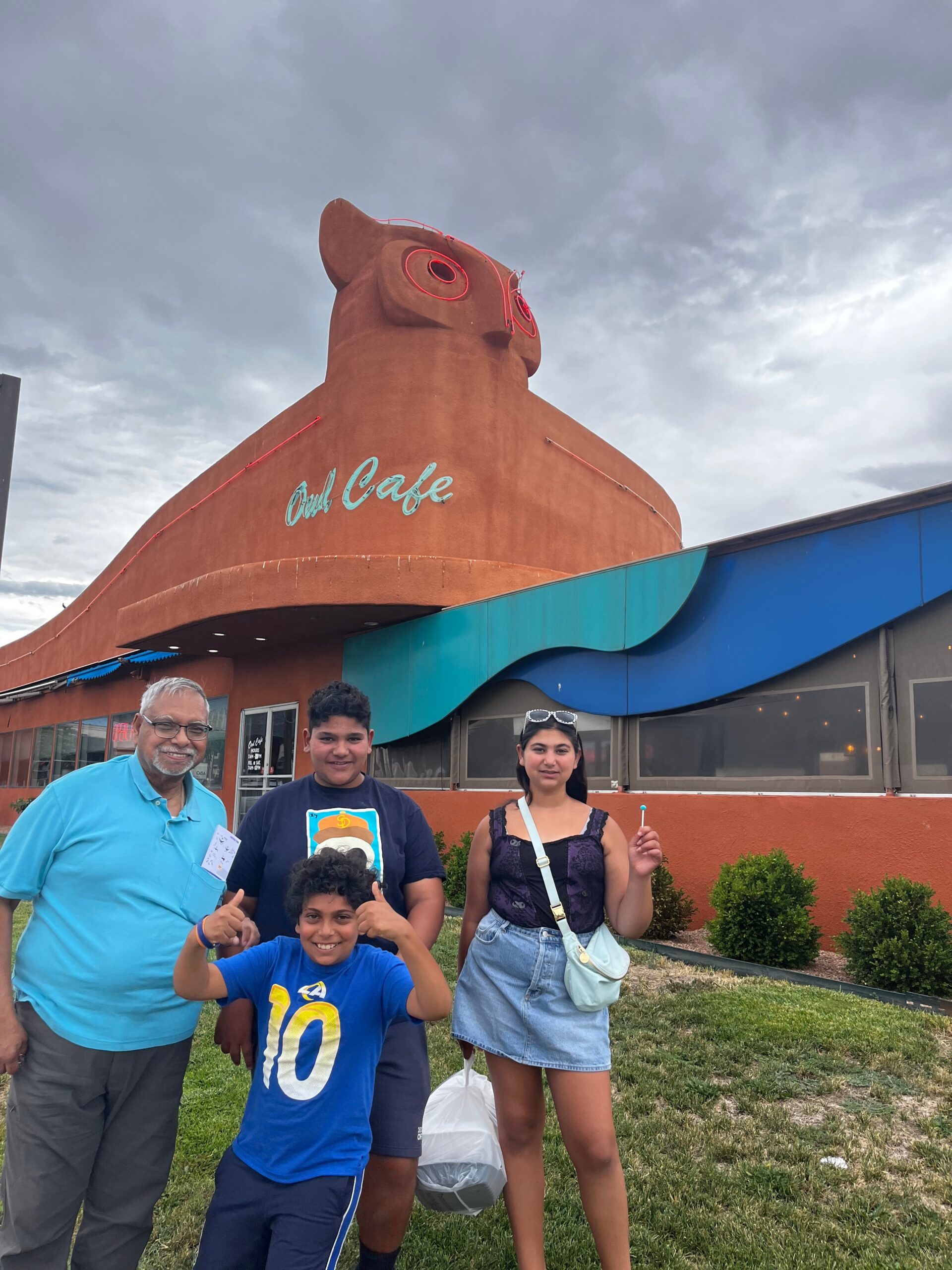 Group of four people posing in front of an orange owl-shaped building with a turquoise sign reading 'Owl Cafe' in a parking lot setting.