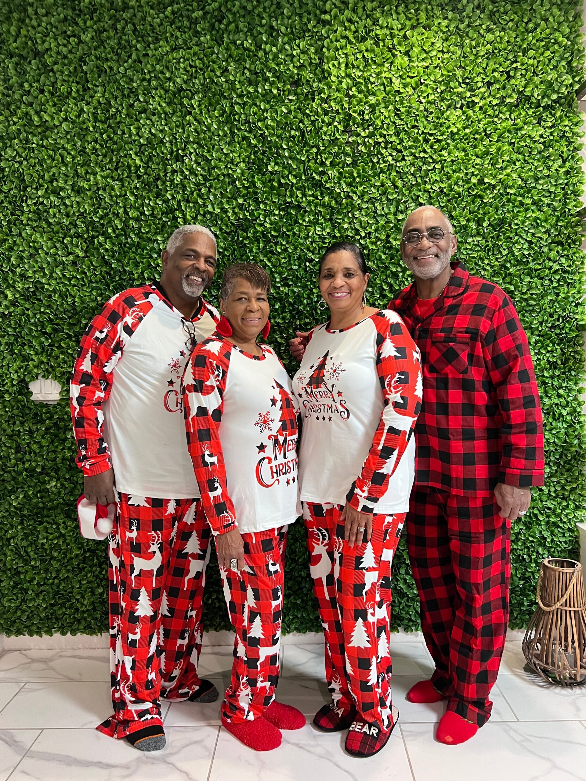 Four smiling adults in Christmas-themed pajamas pose together in front of a green leafy wall.