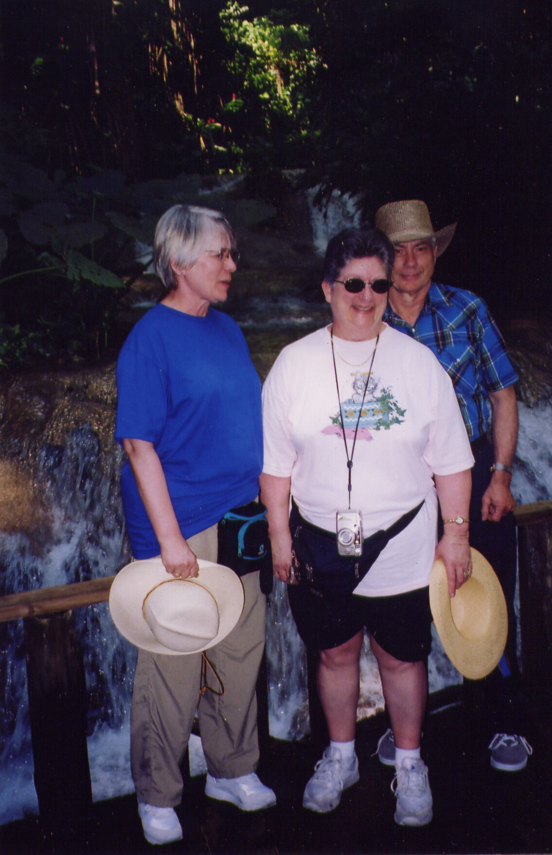 Three people stand together by a small waterfall in a forest, smiling at the camera; two carry wide-brimmed hats, one wears sunglasses and a camera hanging around the neck.