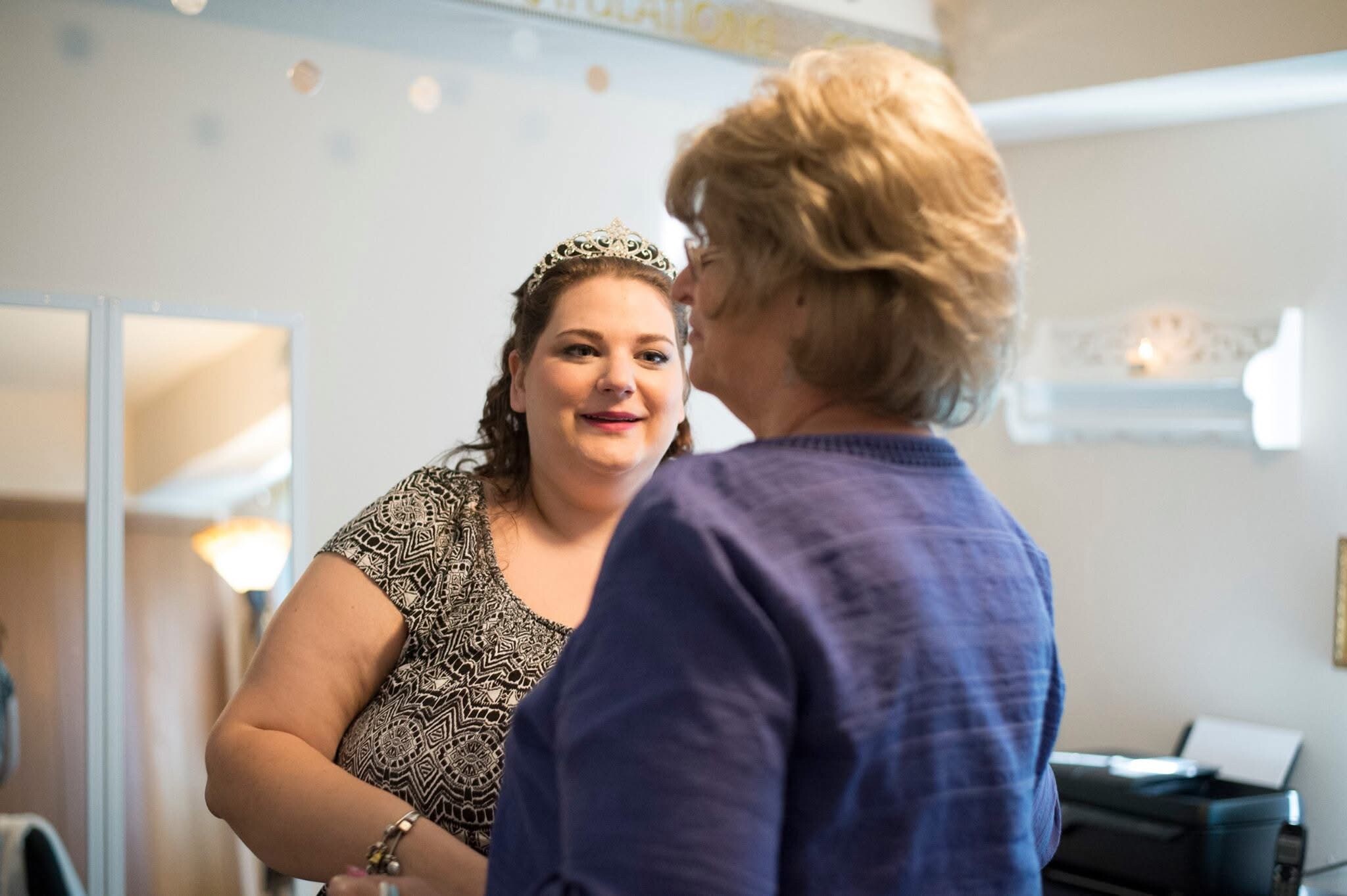 Woman with a decorative tiara and patterned dress smiles at a person in a blue shirt in a dressing room with mirrors nearby.