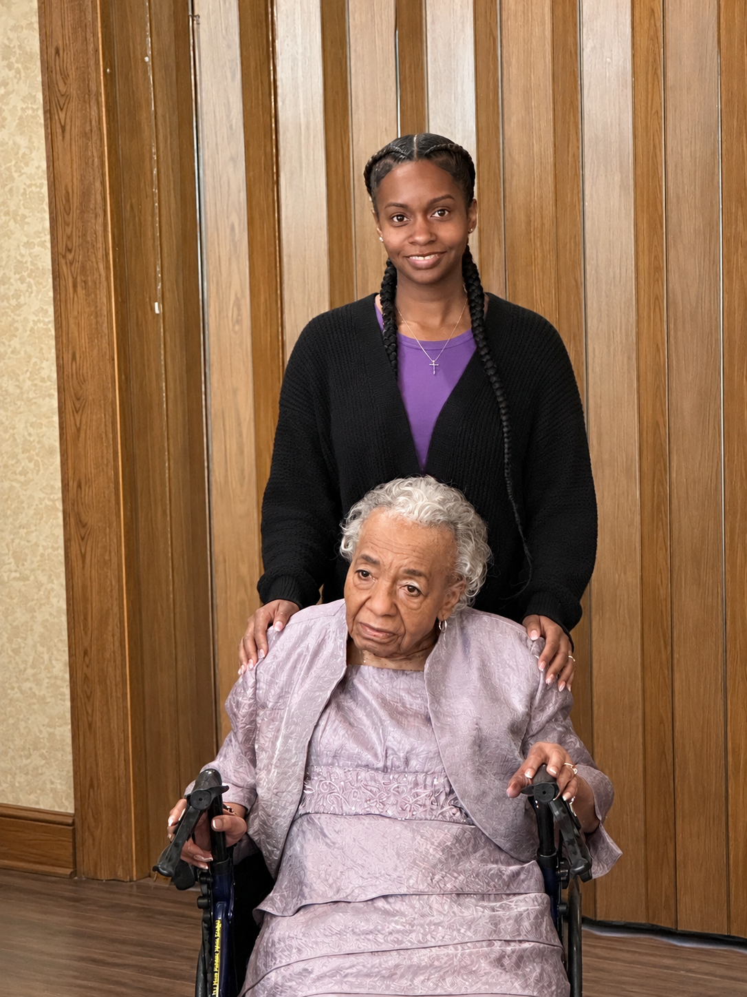 Young woman stands behind an elderly woman in a wheelchair, both looking at the camera in a wood-paneled room.