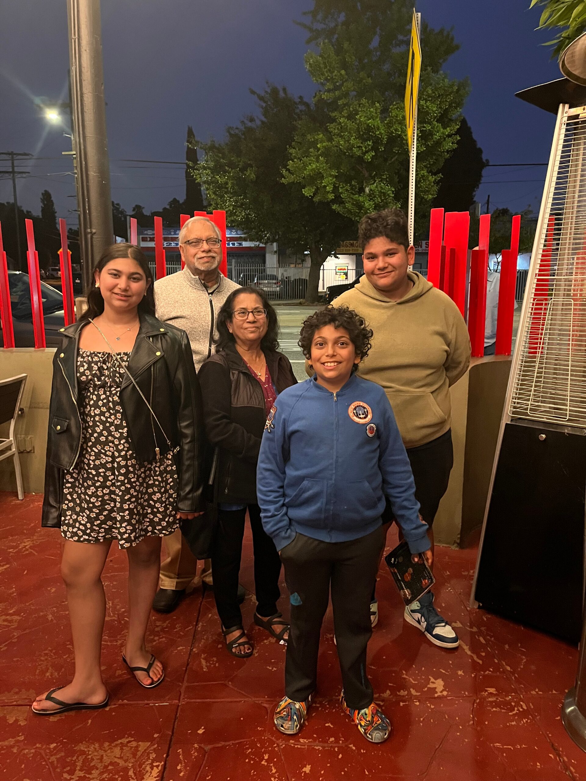 Group of five smiling for a photo outdoors at night, with red vertical sculpture poles behind them and a tall patio heater to the right.