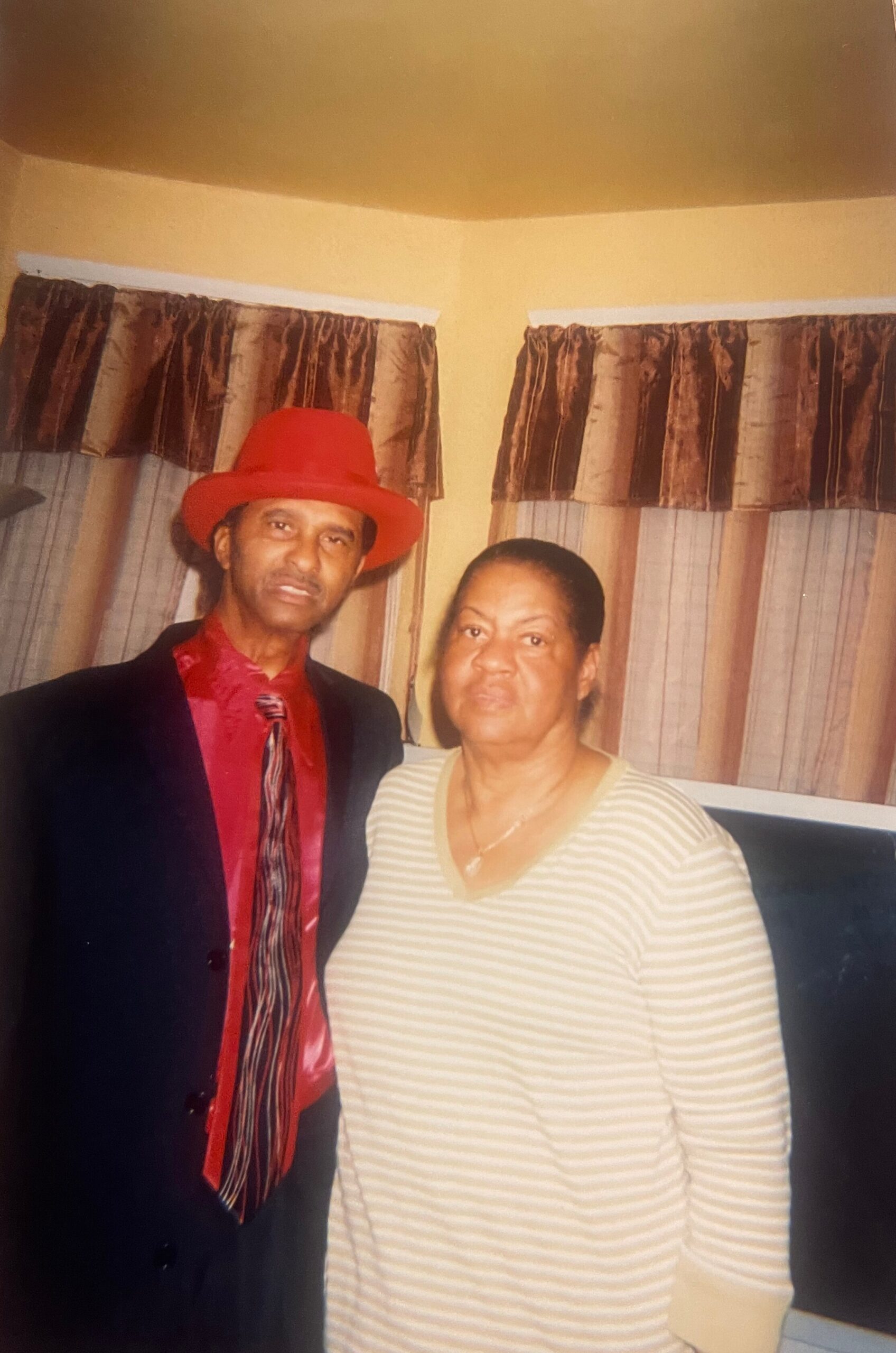 Two people posing indoors: a man in a red hat and suit beside a woman in a striped top, yellow walls behind.