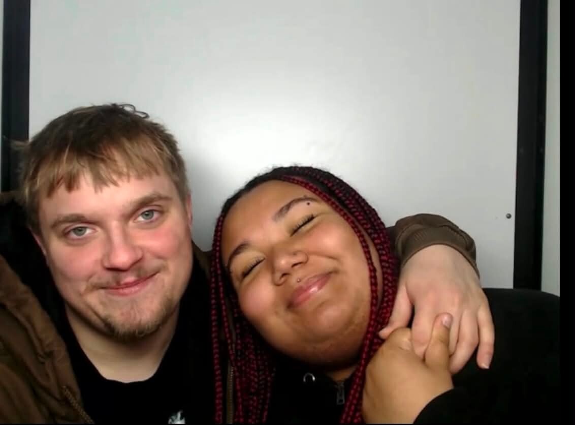 Two friends posing for a selfie: a light-skinned man with short blond hair smiles as he wraps his arm around a smiling woman with red braids.