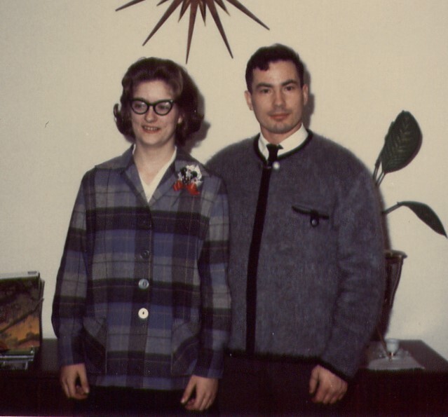 Couple posing for a vintage indoor photo: woman in a plaid coat with glasses and a corsage, man in a gray sweater beside her.