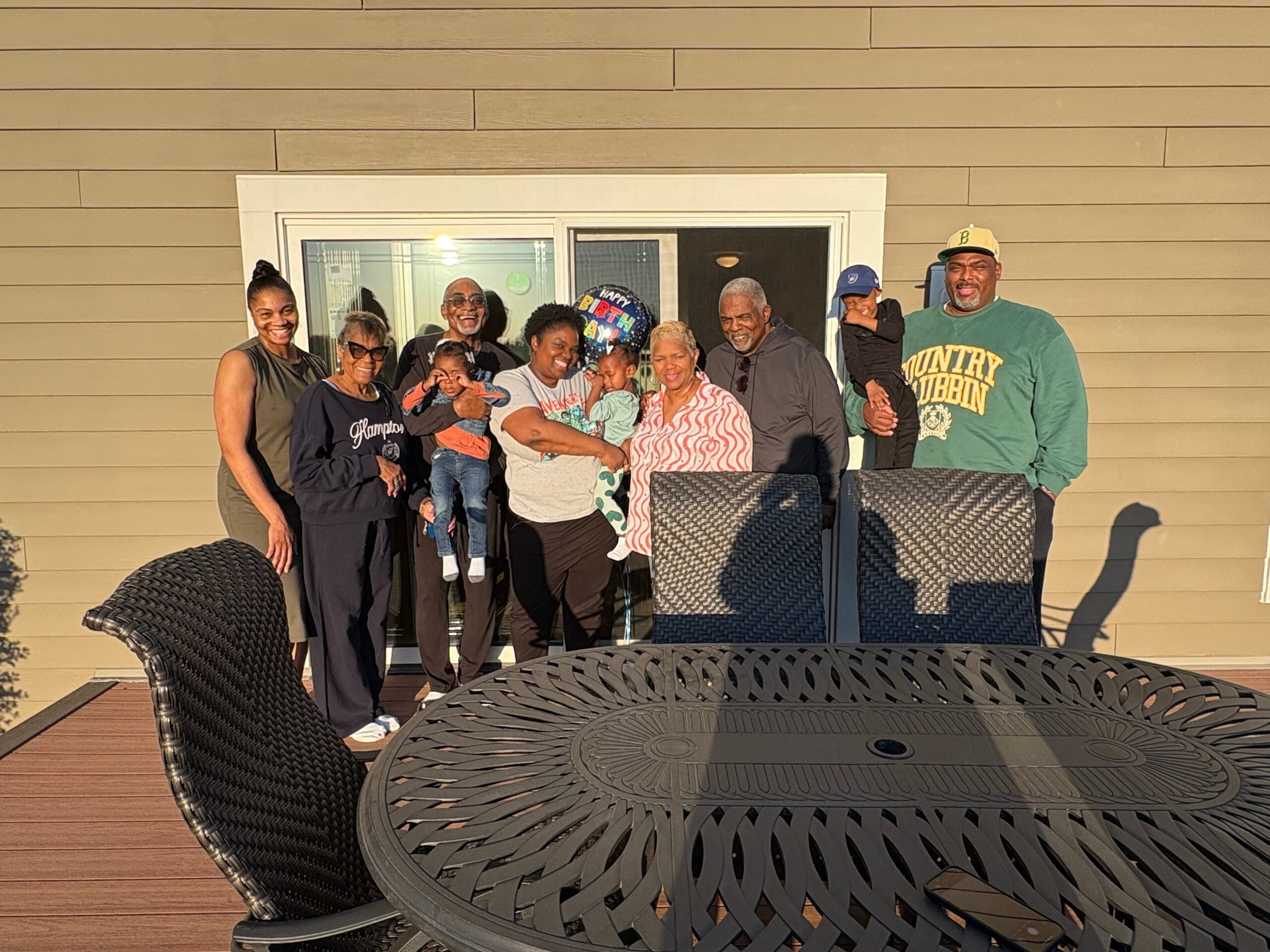 A multigenerational family posing on a wooden deck outside a house, with a birthday balloon in the background and wicker furniture in the foreground.