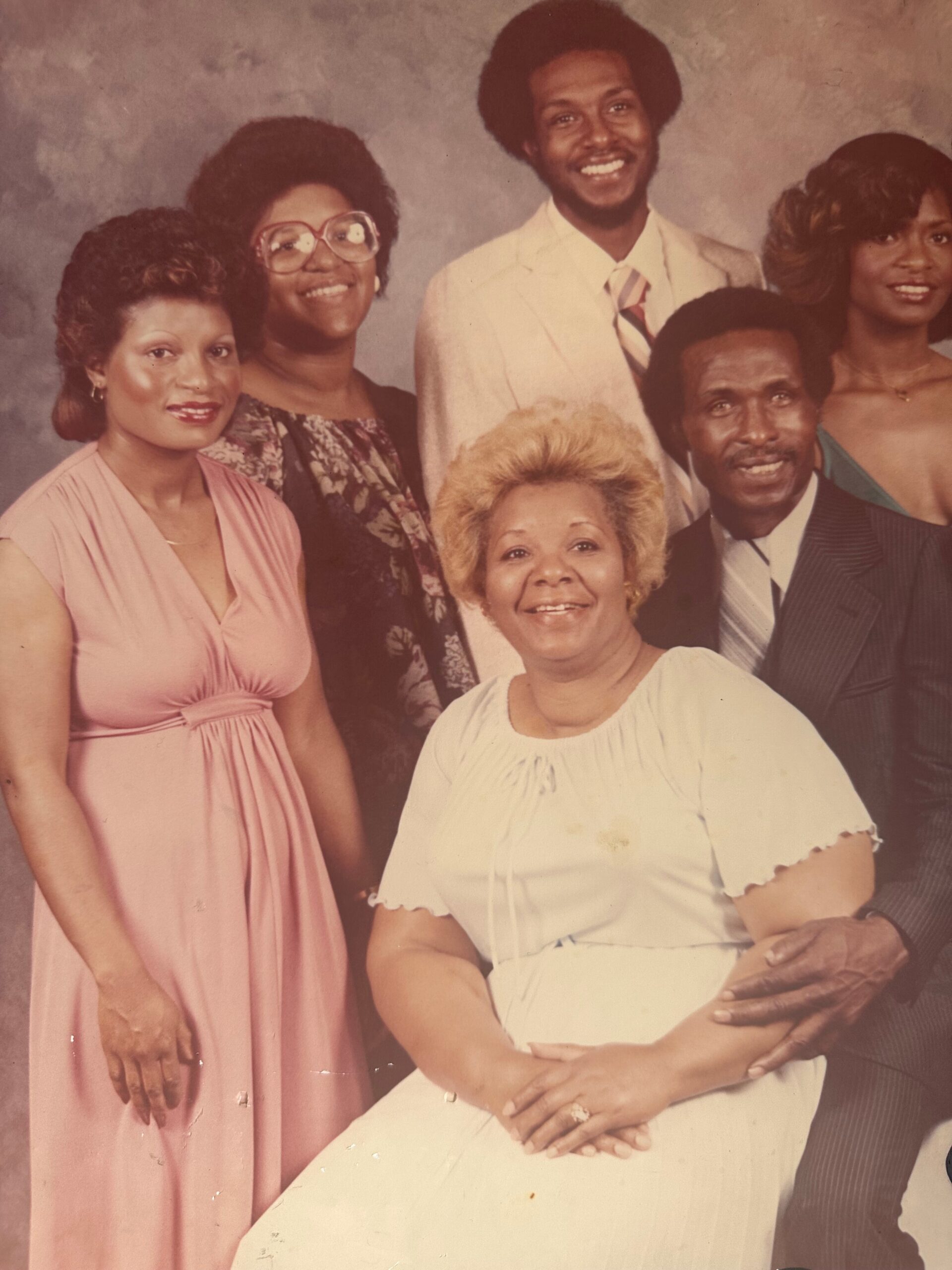 Smiling multigenerational family portrait in formal attire: woman in pink dress left, seated elderly woman in white front, and others standing around.