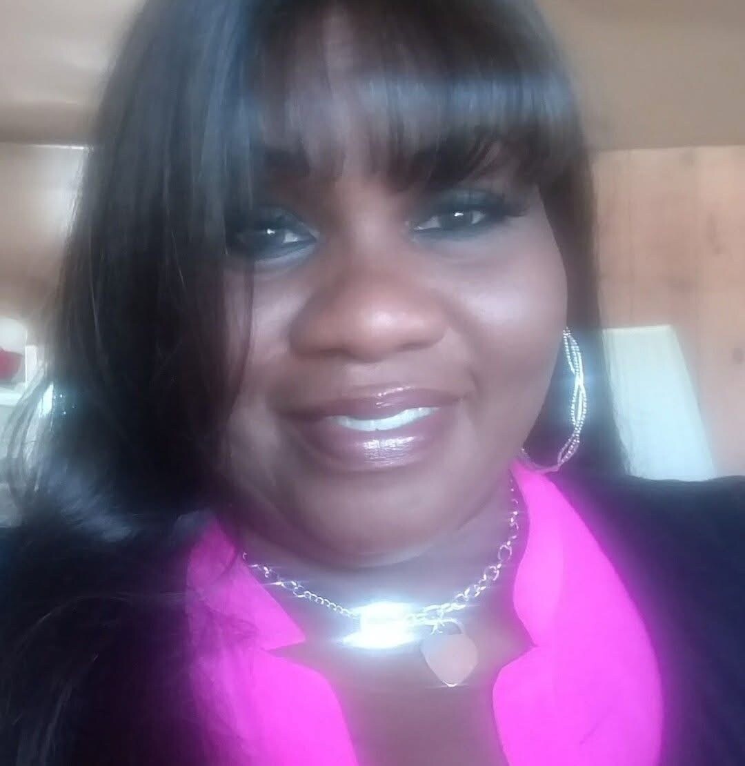 Smiling woman with long dark hair, pink blouse, and silver hoop earrings in an indoor setting.