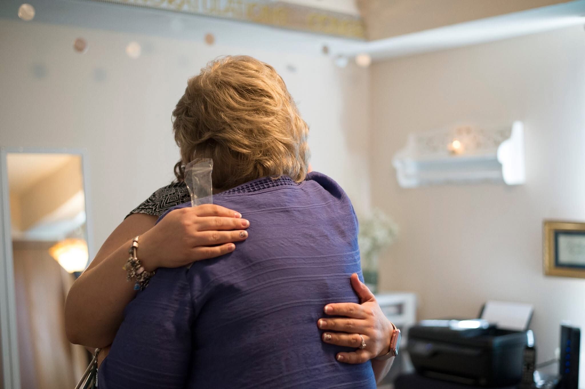Two people share a warm hug, facing away from the camera; one in a purple shirt with patterned sleeves visible on the other person’s arm touches them.