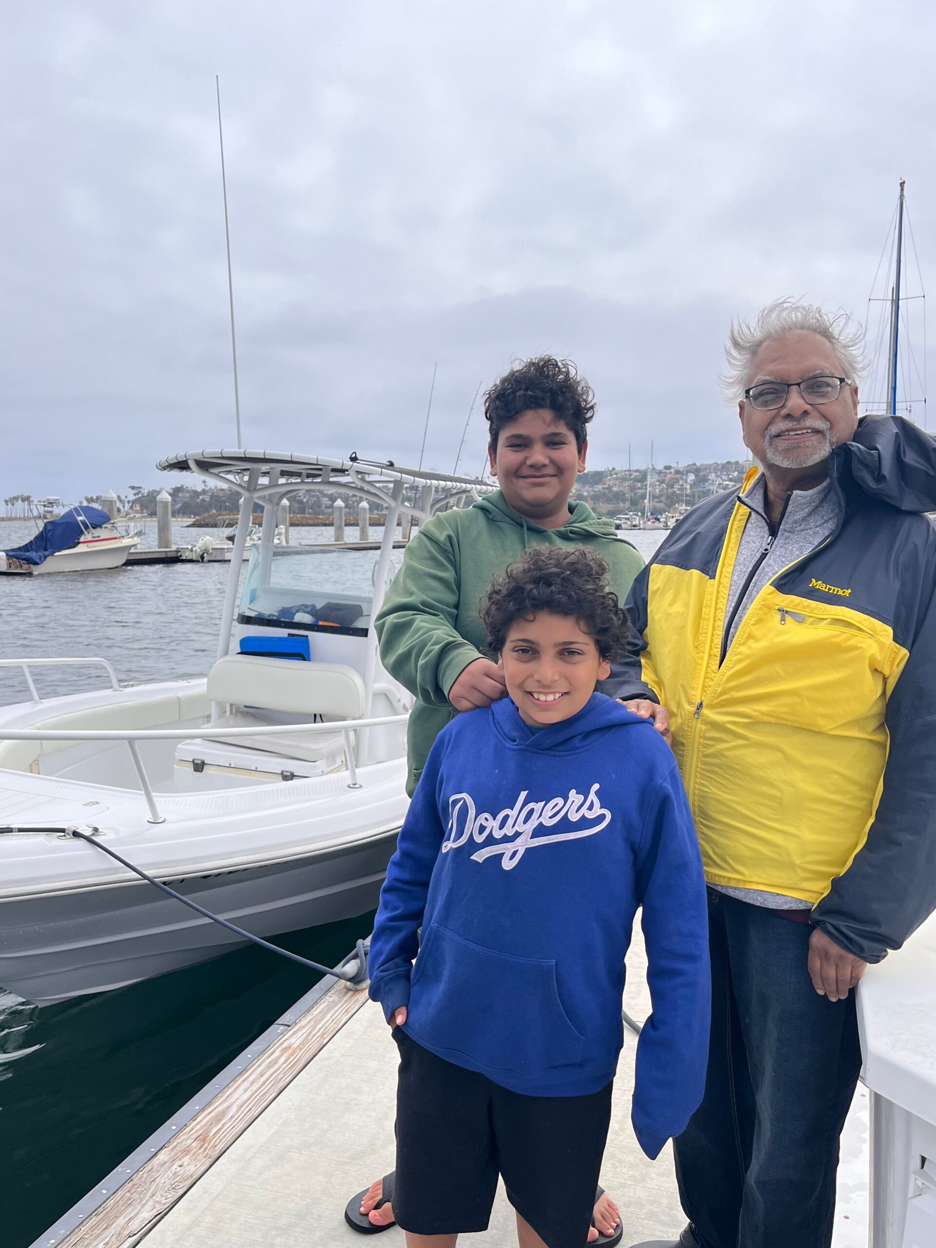 Three people stand on a white boat at a marina, smiling with sailboats and a cloudy sky in the background.