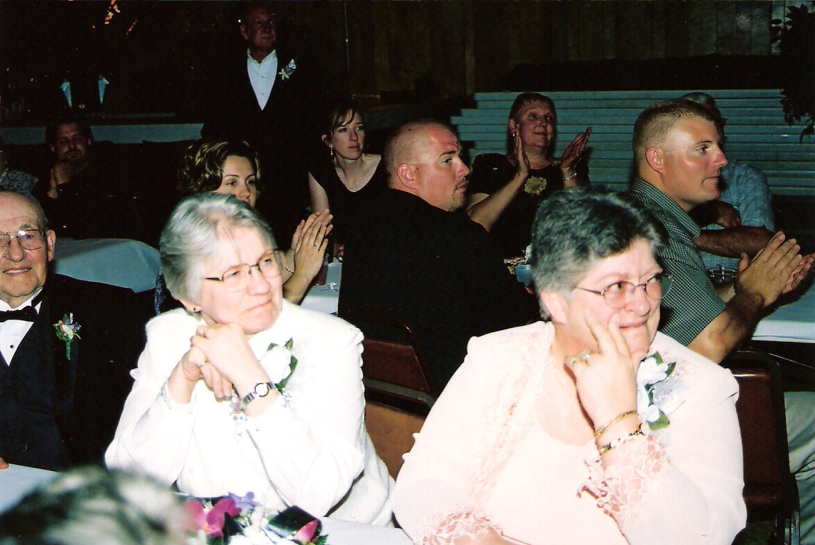Group of guests seated at a wedding reception, two older women in light outfits with corsages in the foreground.