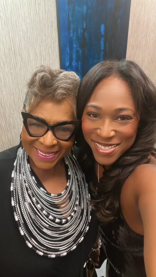 Two women smile for a selfie indoors; left woman wears bold black cat-eye glasses and layered black-and-white necklace, right woman in a sleeveless black dress.