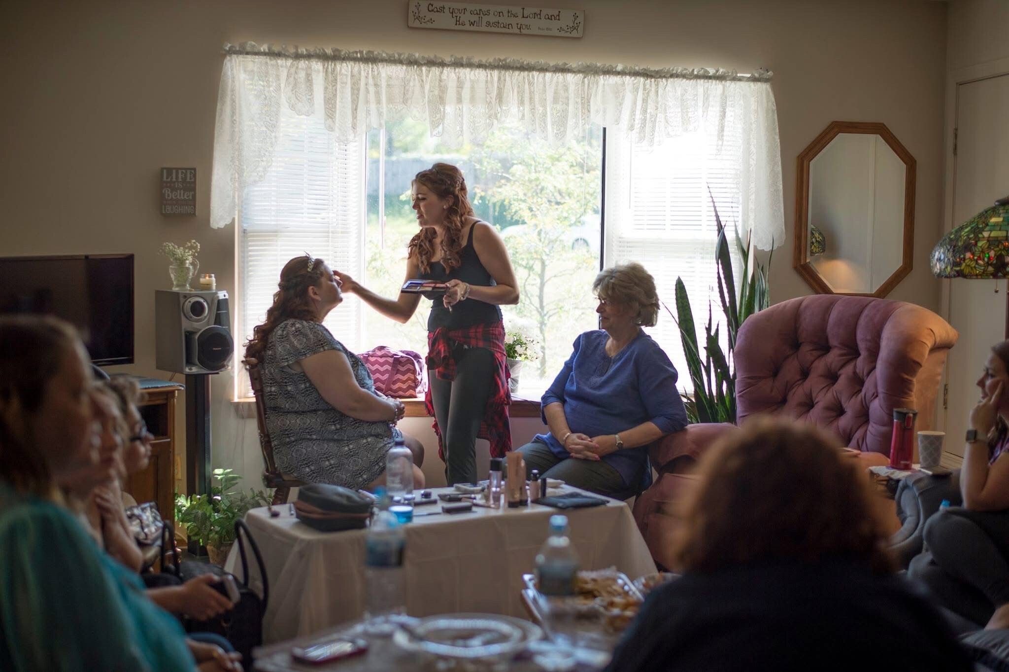 Woman applying makeup to seated friend while others watch in a cozy living room. A makeup session around a table with snacks and drinks nearby.