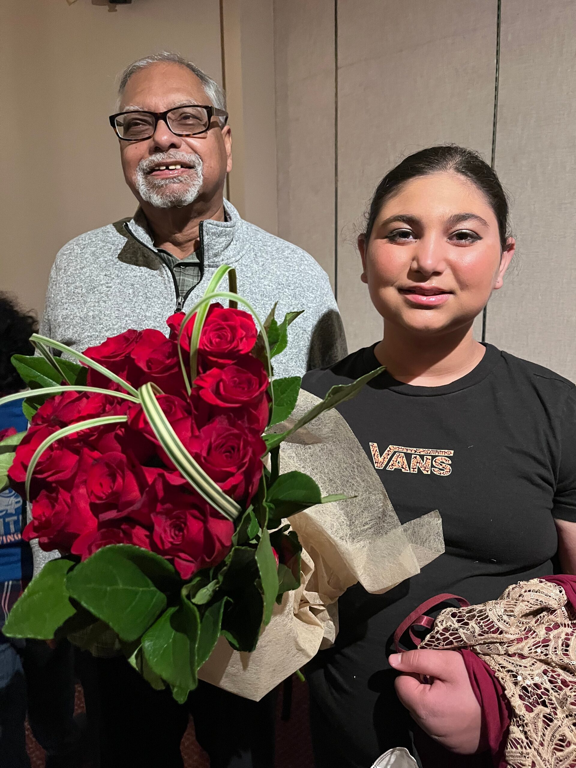 Older man with glasses smiles while holding a bouquet of red roses, standing beside a young woman in a dark shirt.