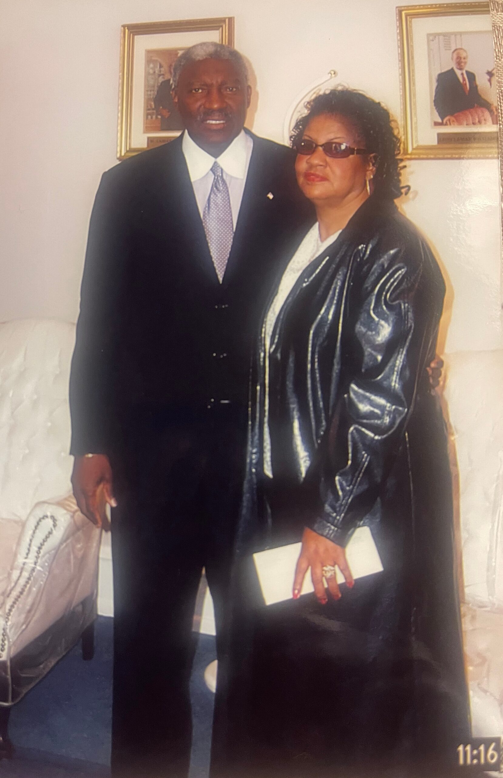 Two adults stand side by side in a living room; man in a dark suit and tie, woman in a shiny black coat with sunglasses.