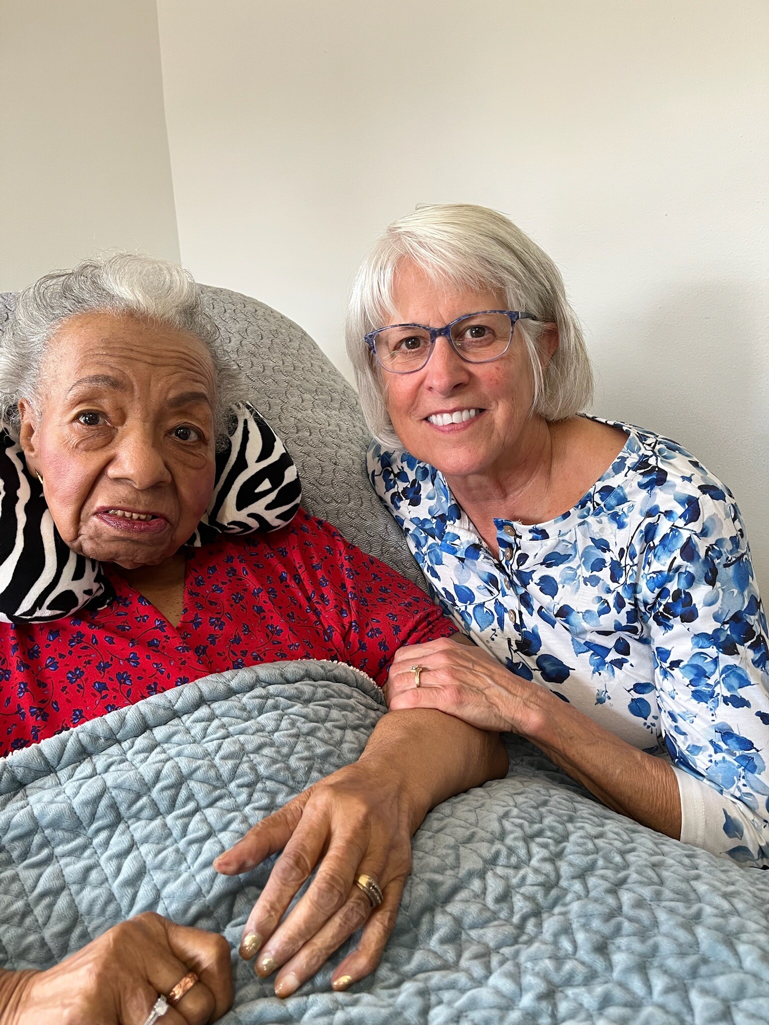 Two elderly women sit close together on a bed, holding hands and smiling at the camera.