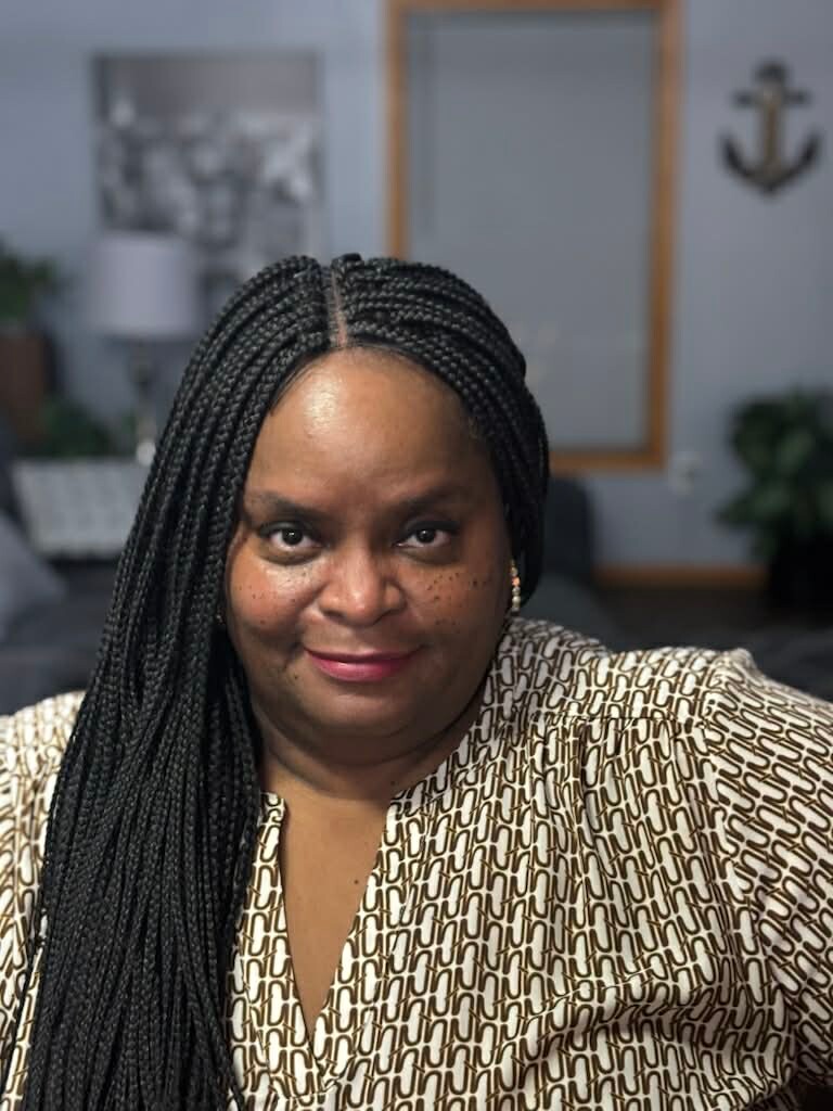 Portrait of a smiling woman with long braided hair in a patterned blouse, indoors in a living room setting.