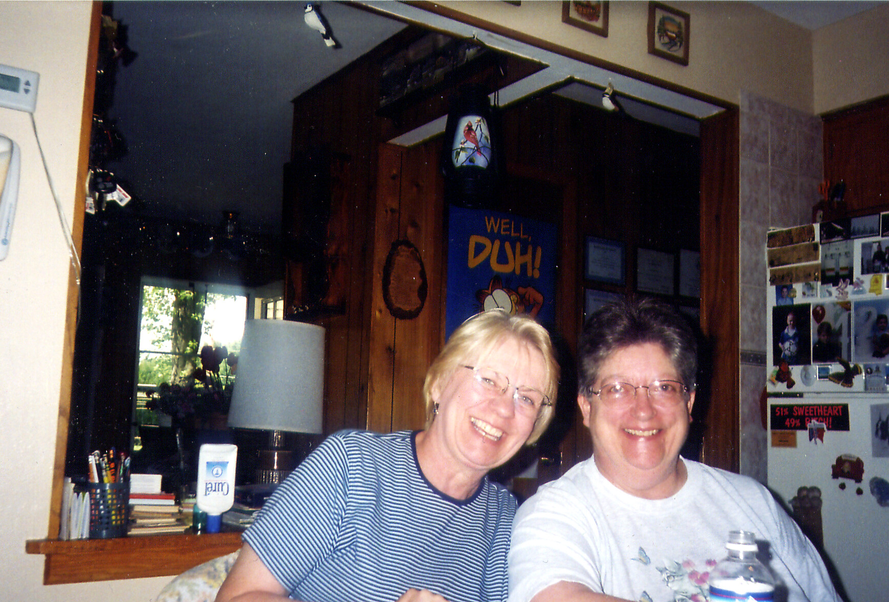 Two smiling women posing together at a kitchen counter, with photos on the fridge behind them and a blue poster reading 'WELL DUH!' in the background.