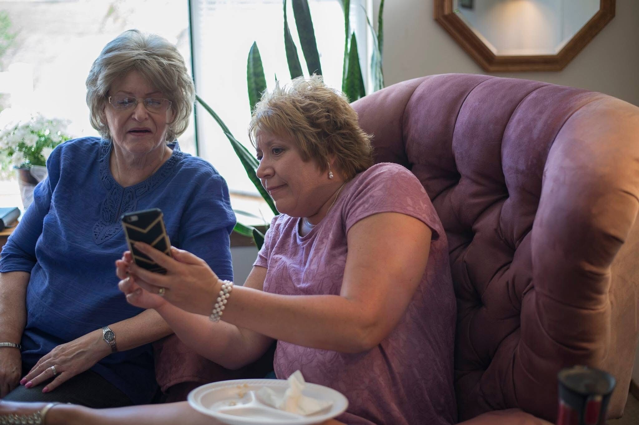 Two elderly women sit on a purple couch, looking at a smartphone together inside a living room.