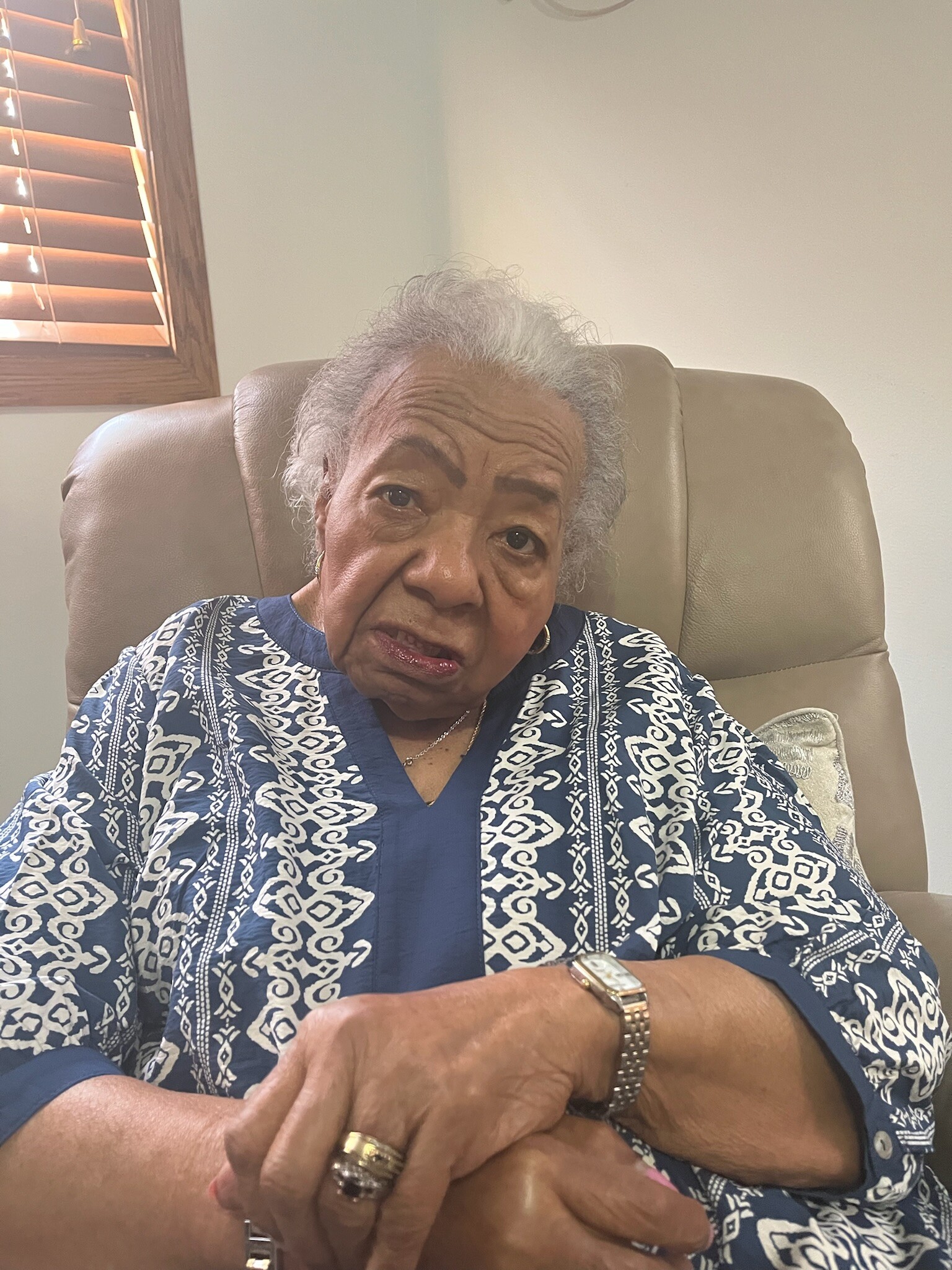 Close-up of an elderly woman with gray hair, wearing a blue patterned blouse, seated in a beige chair with hands resting on a table.