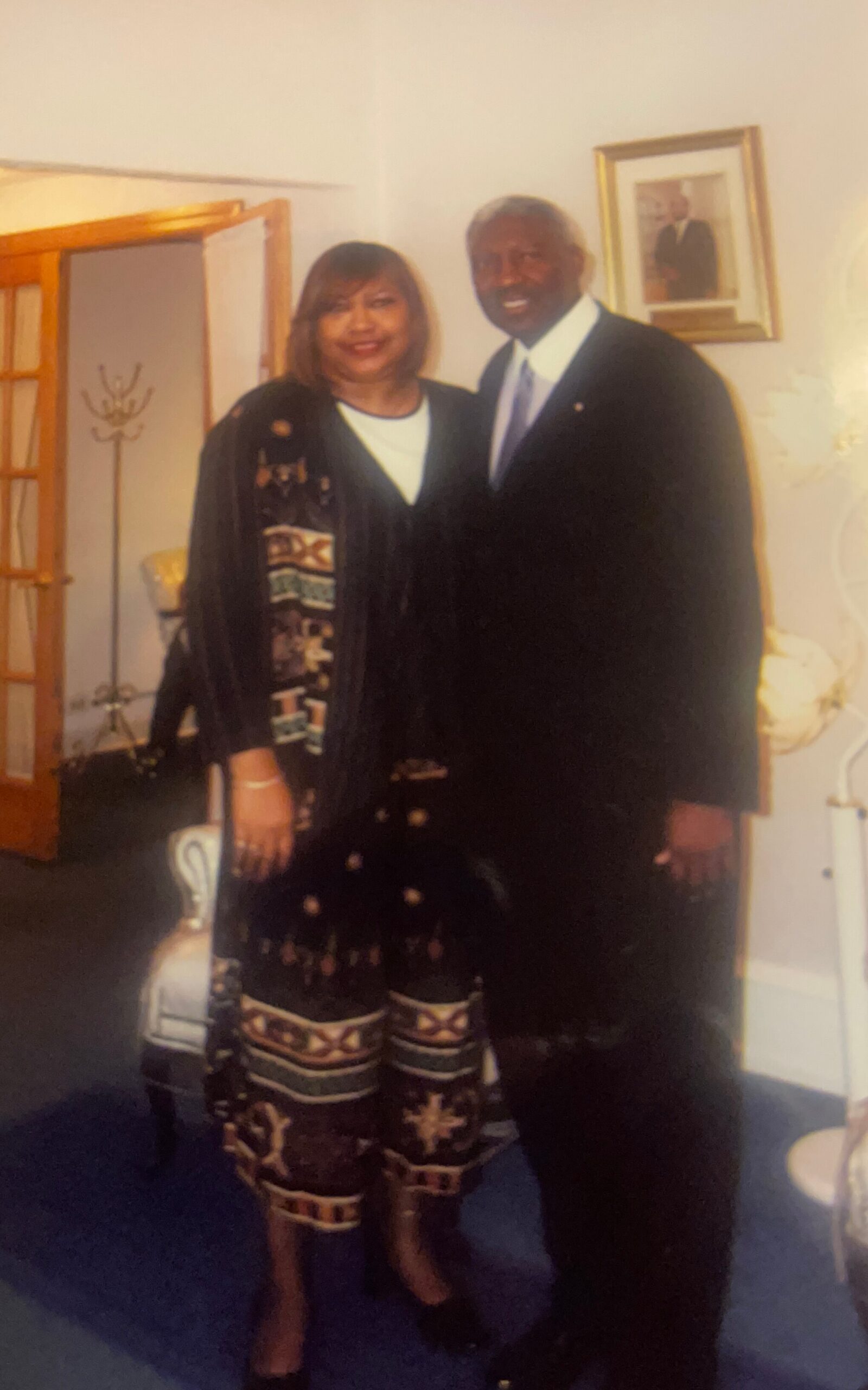 Smiling couple posing together in a living room, woman in a patterned black outfit and man in a dark suit.