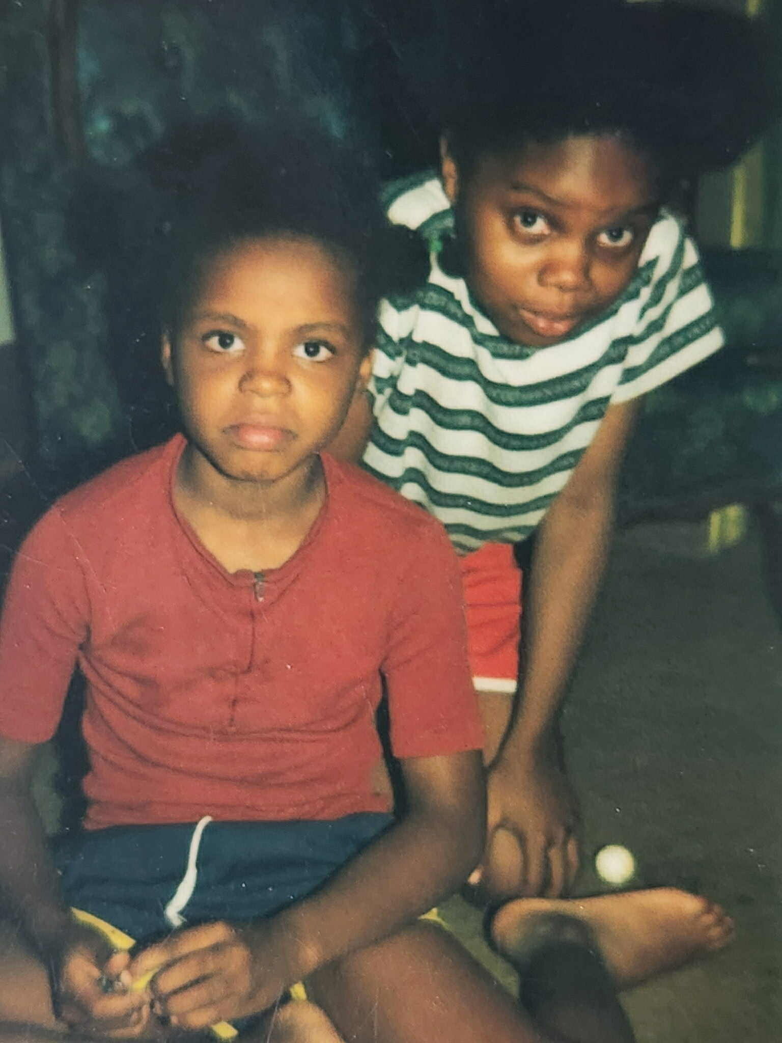 Two young children sit close together; a girl in a striped shirt looks toward the camera while a boy in a red shirt sits beside her, both holding a toy nearby.