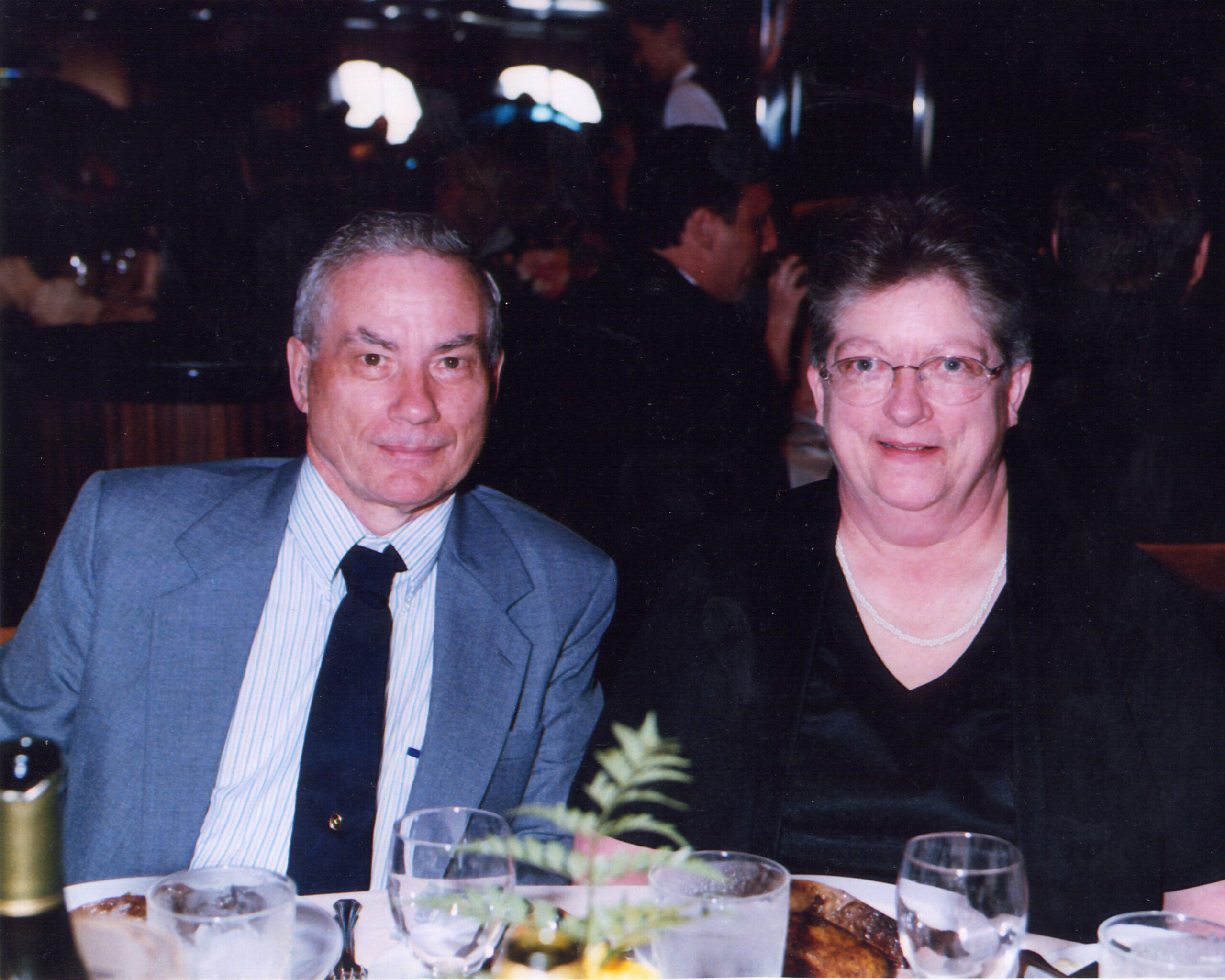 A smiling older couple sits at a dimly lit dinner table with wine glasses and assorted dishes in front of them.