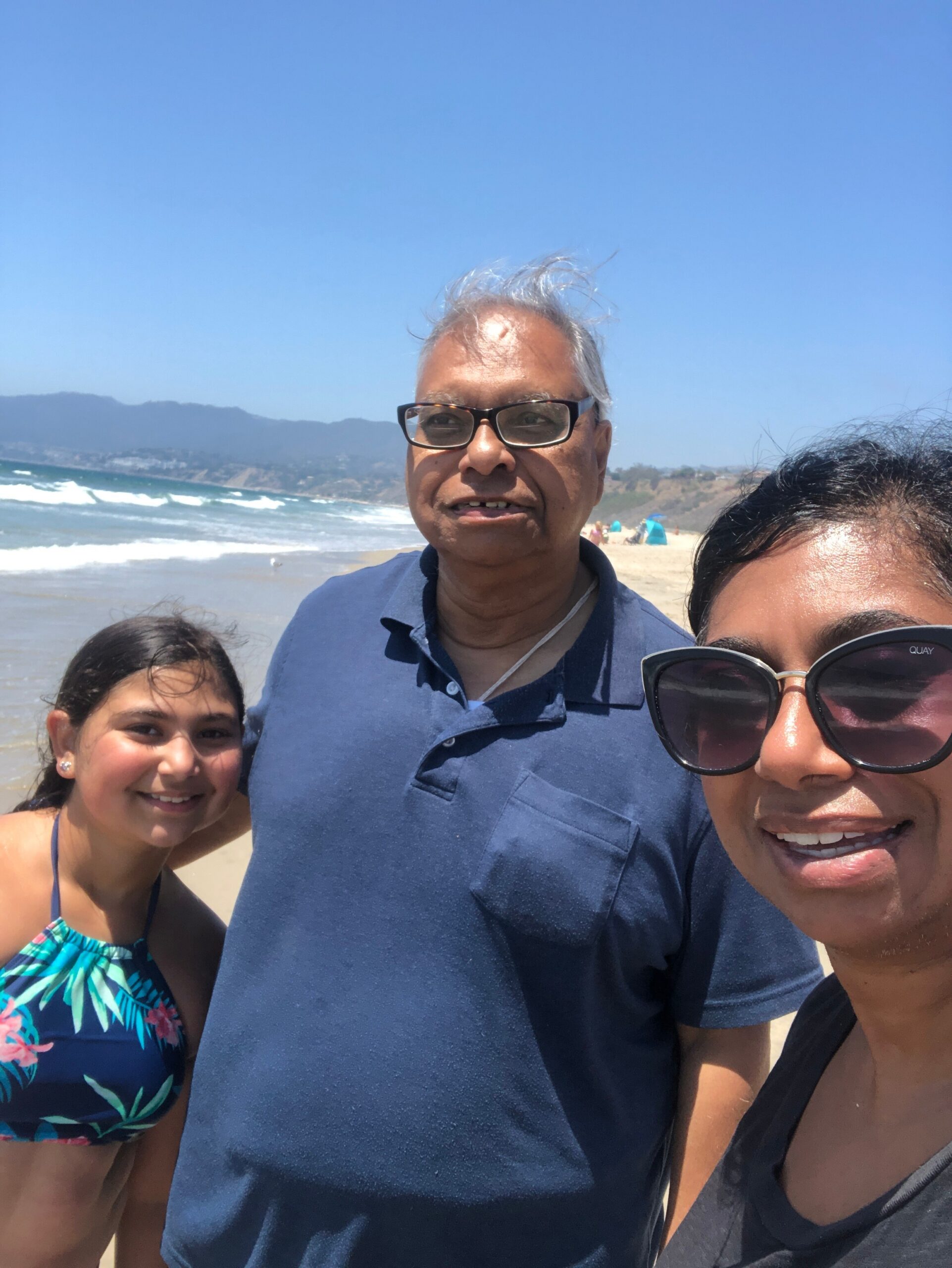 Family selfie at the beach with ocean and distant mountains in the background; three people smiling in foreground (man with glasses center, woman on right wearing sunglasses, girl on left).