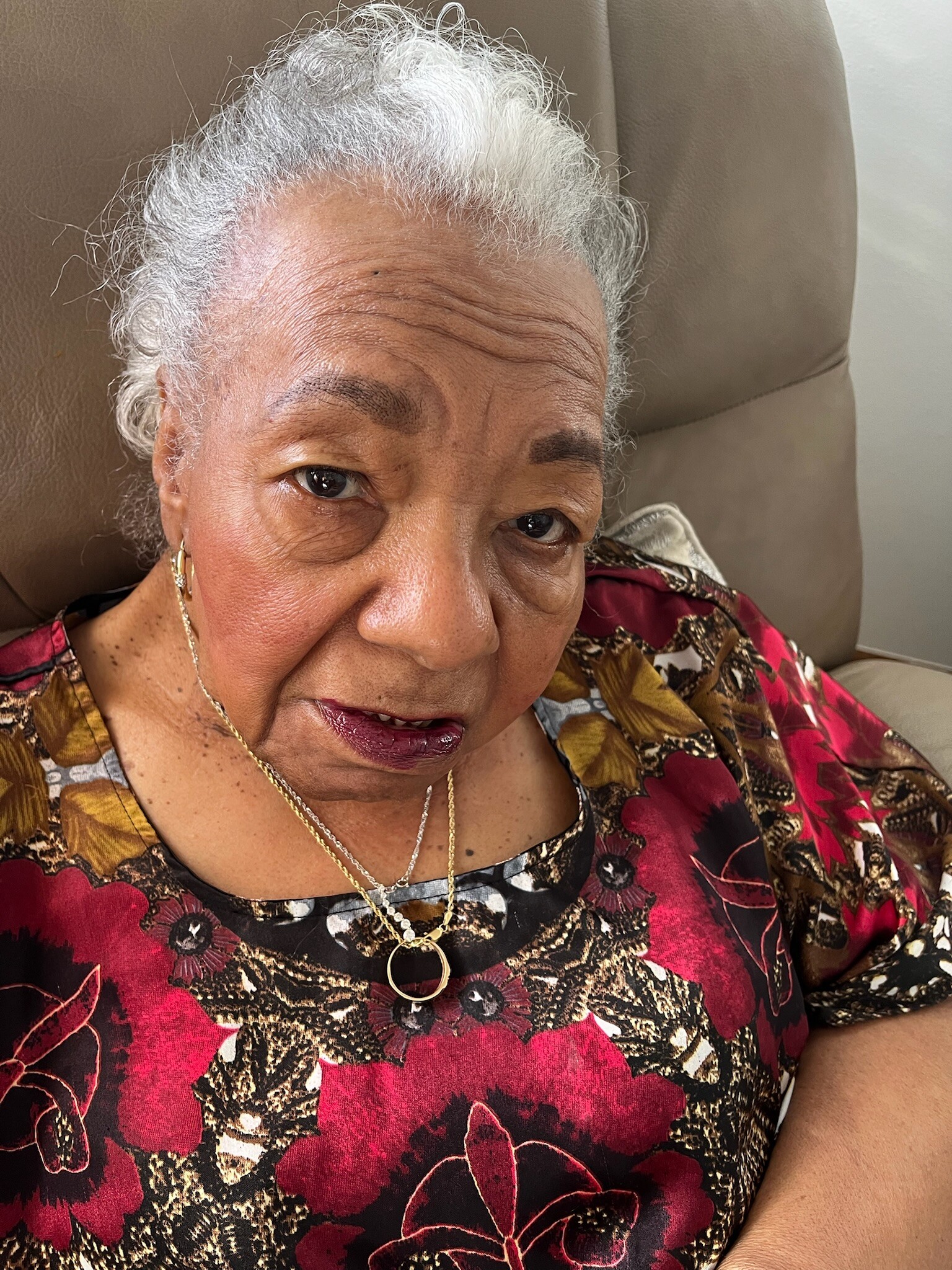 Close-up of an elderly woman with short white hair wearing a red and black floral blouse, gold necklace, seated on a tan chair.